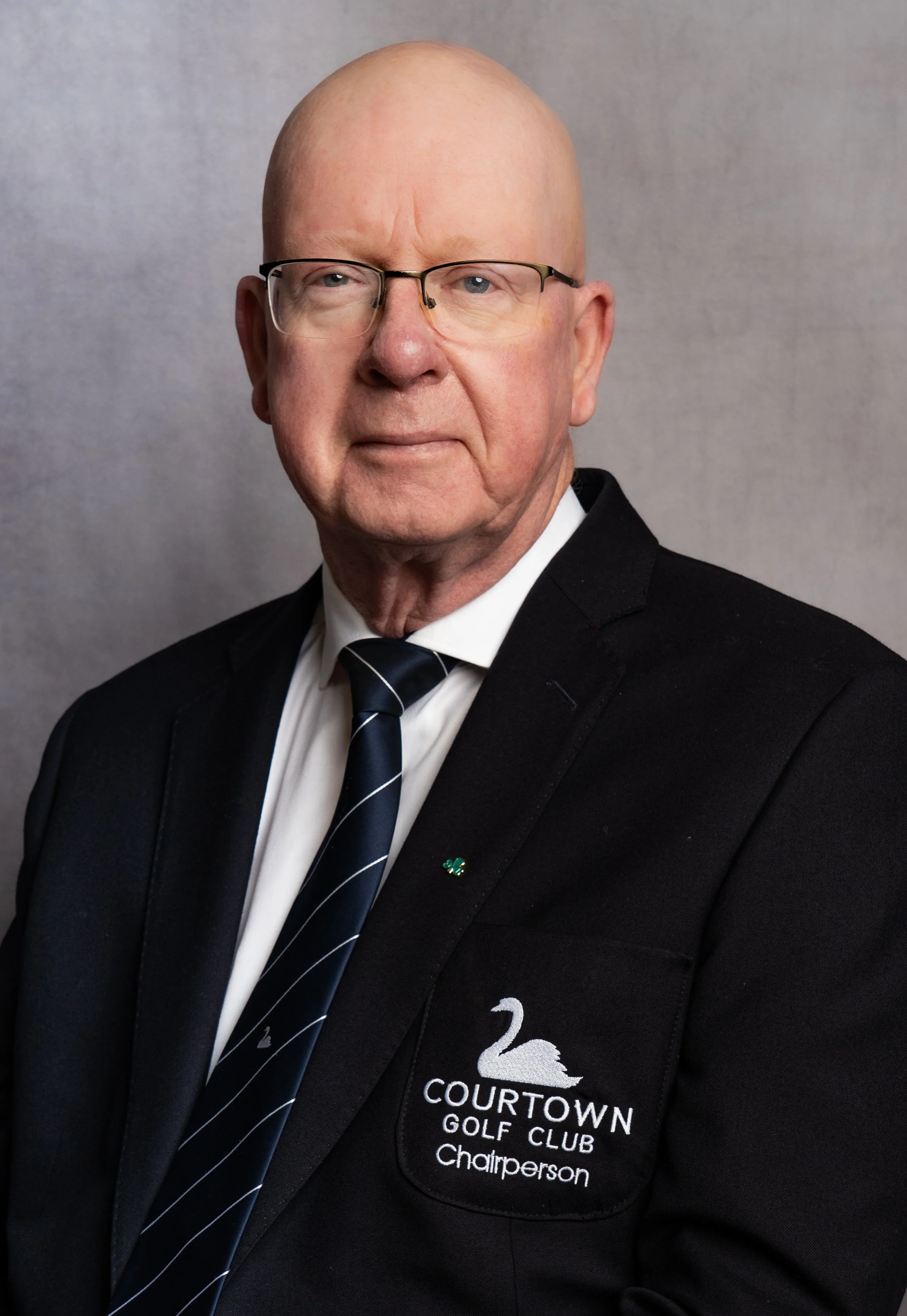 An elderly man in a black suit with a 'Courtown Golf Club' logo and the word 'Chairperson' on it, wearing glasses and a striped tie, standing against a plain gray background.
