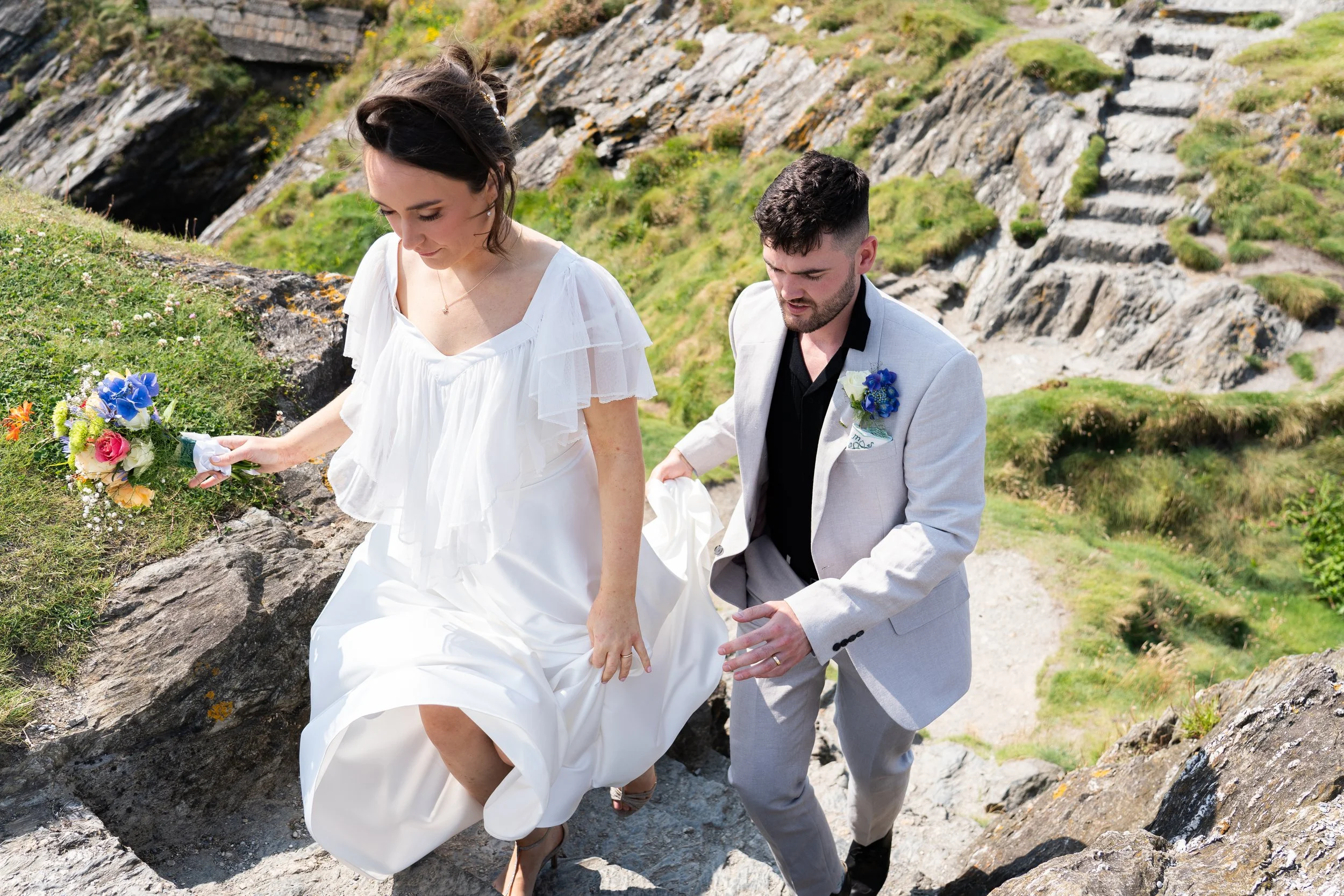 A couple in wedding attire climbing a rocky outdoor trail with green hills and rocks in the background. The bride holds a bouquet of colorful flowers, and the groom helps her as they ascend the trail.