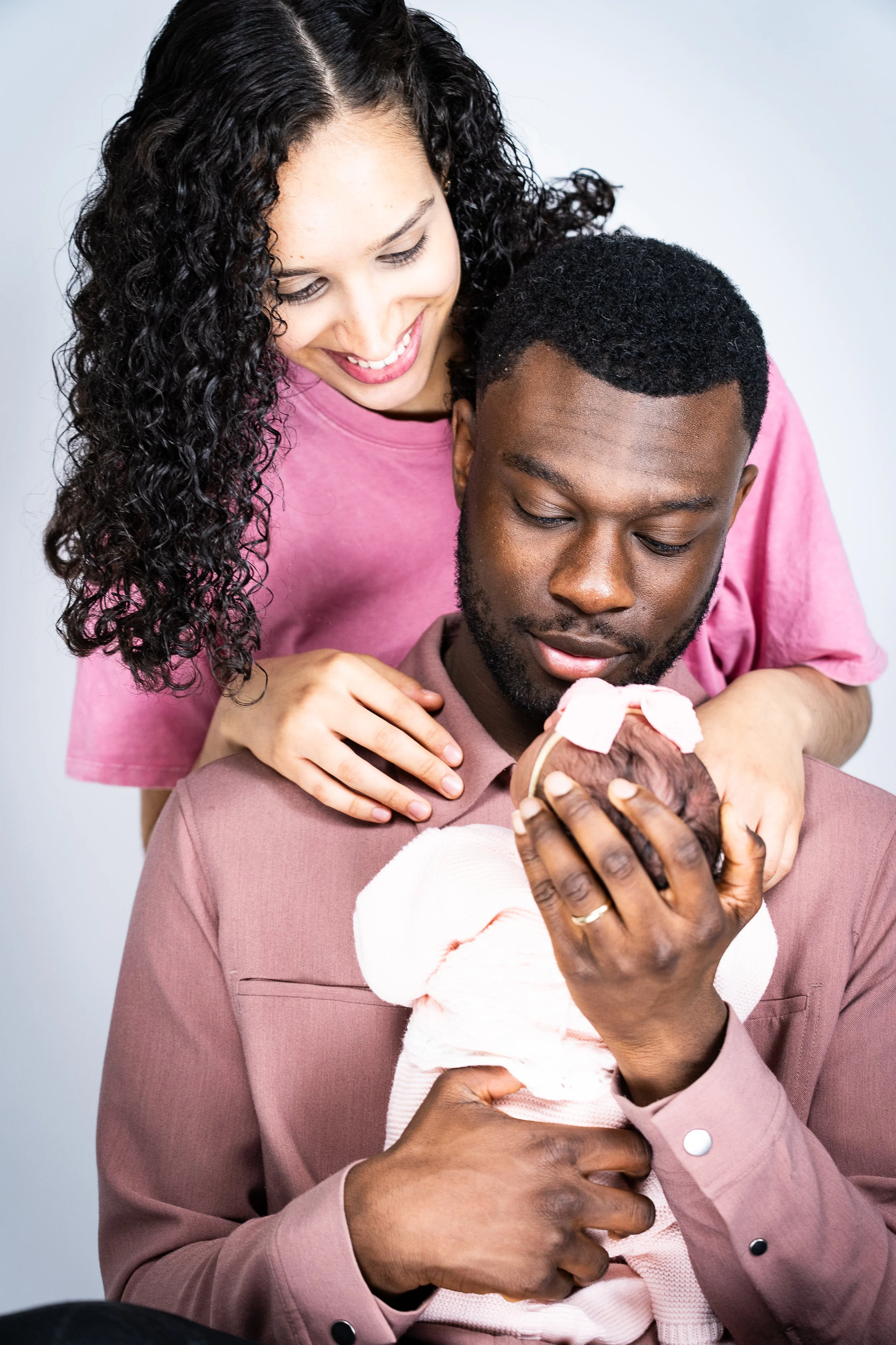 A woman lovingly looking at a newborn baby wrapped in a pink blanket, with her hand resting on the man's shoulder.