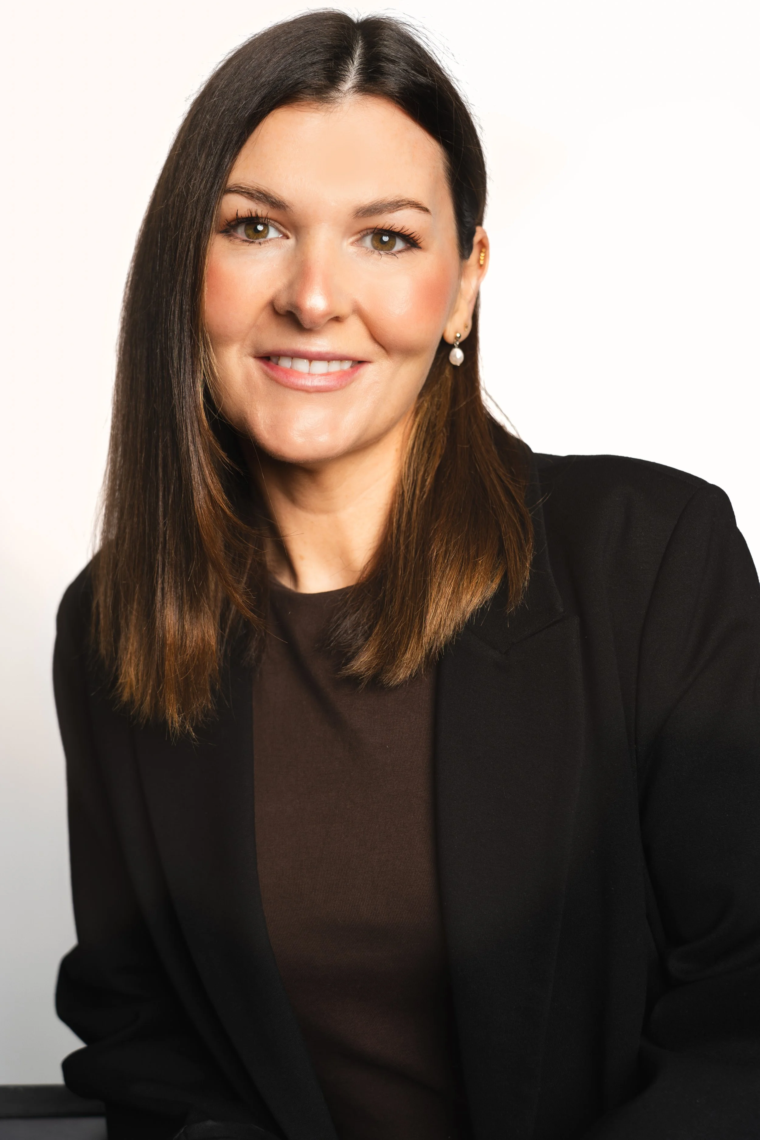 A professional woman with shoulder-length brown hair, light skin, wearing a black blazer and brown top, smiling against a plain white background.