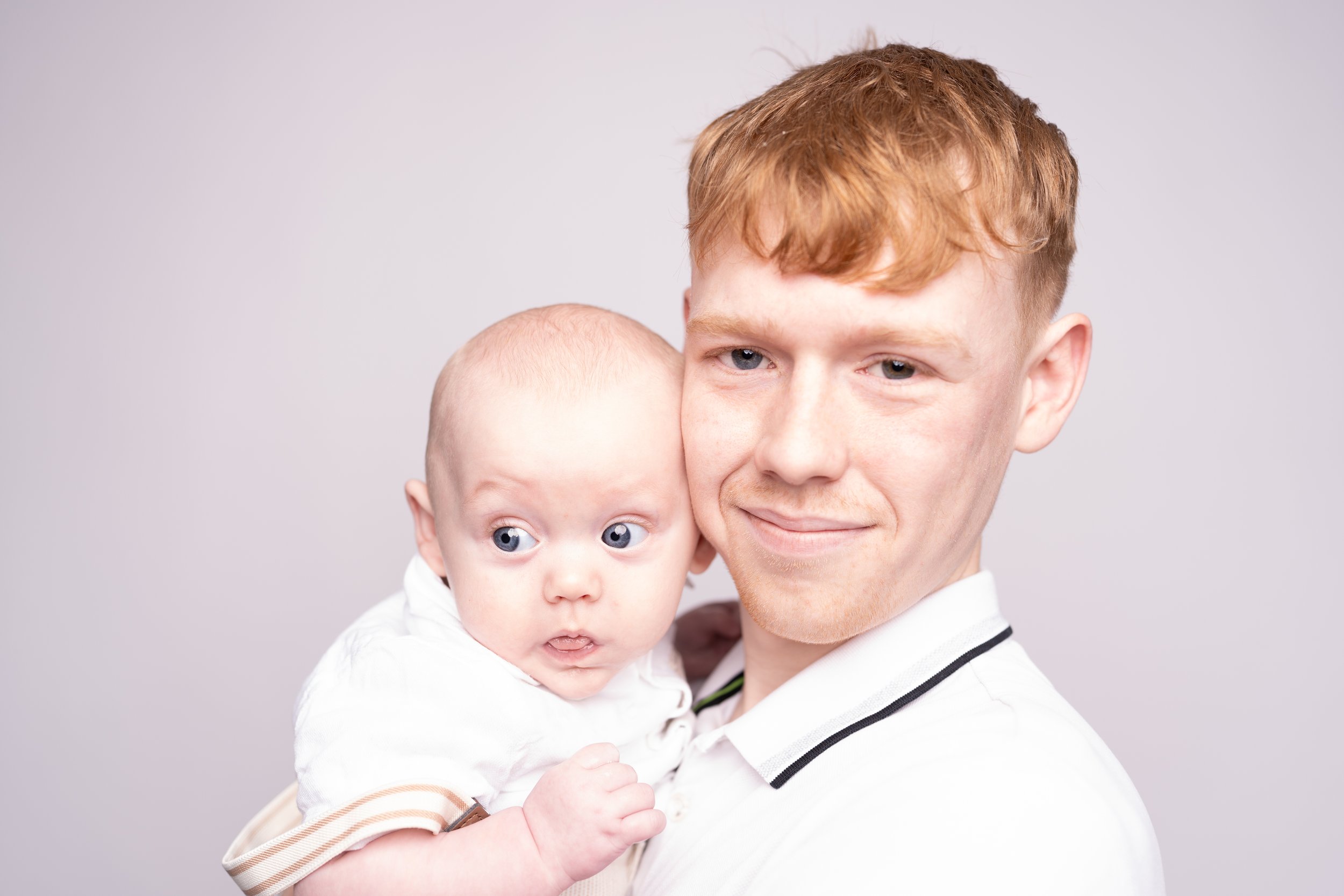 A young man holding a baby with light skin tone and red hair, both looking at the camera against a plain light gray background.