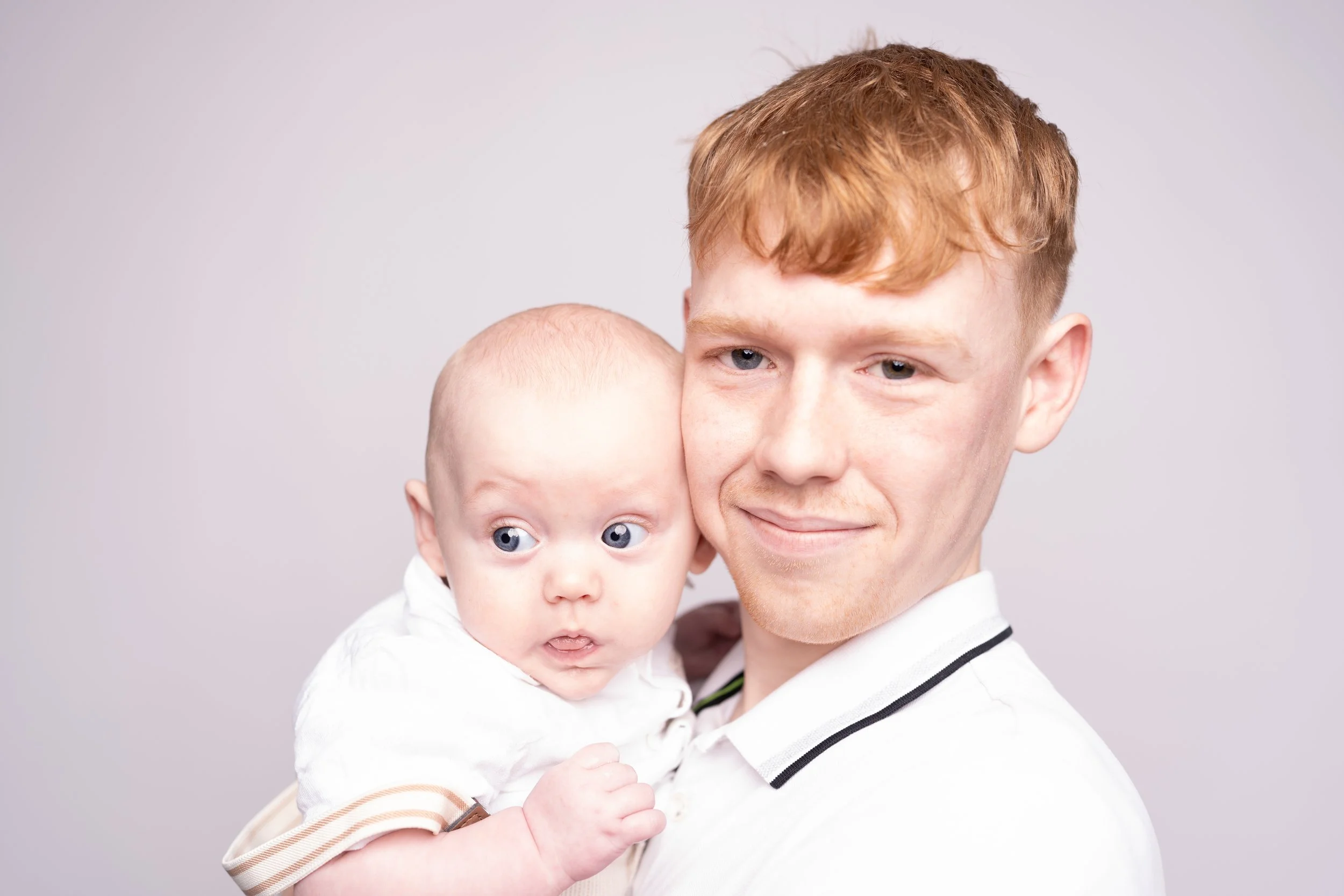 A young man holding a baby with light skin tone and red hair, both looking at the camera against a plain light gray background.