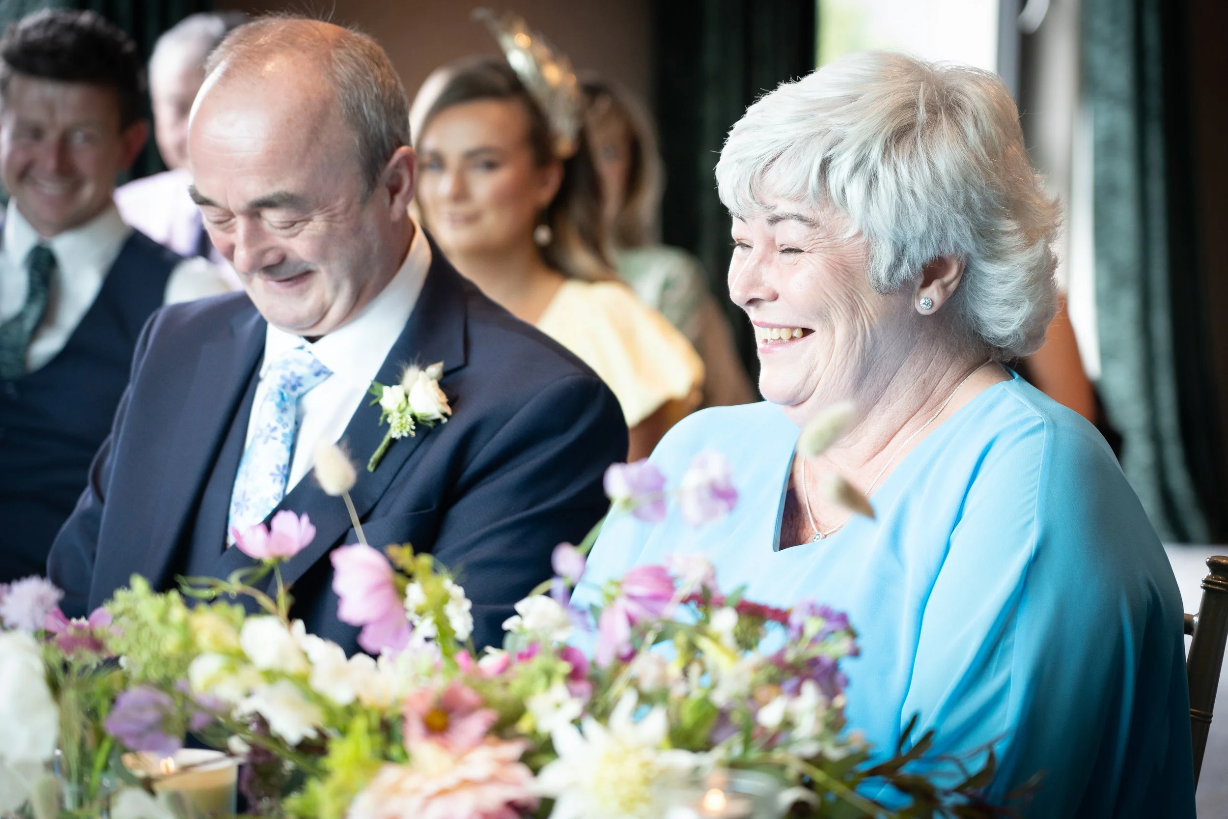 A group of people smiling and laughing at a wedding reception, with a focus on an elderly woman with white hair in a blue dress and a middle-aged man in a suit, surrounded by flowers.