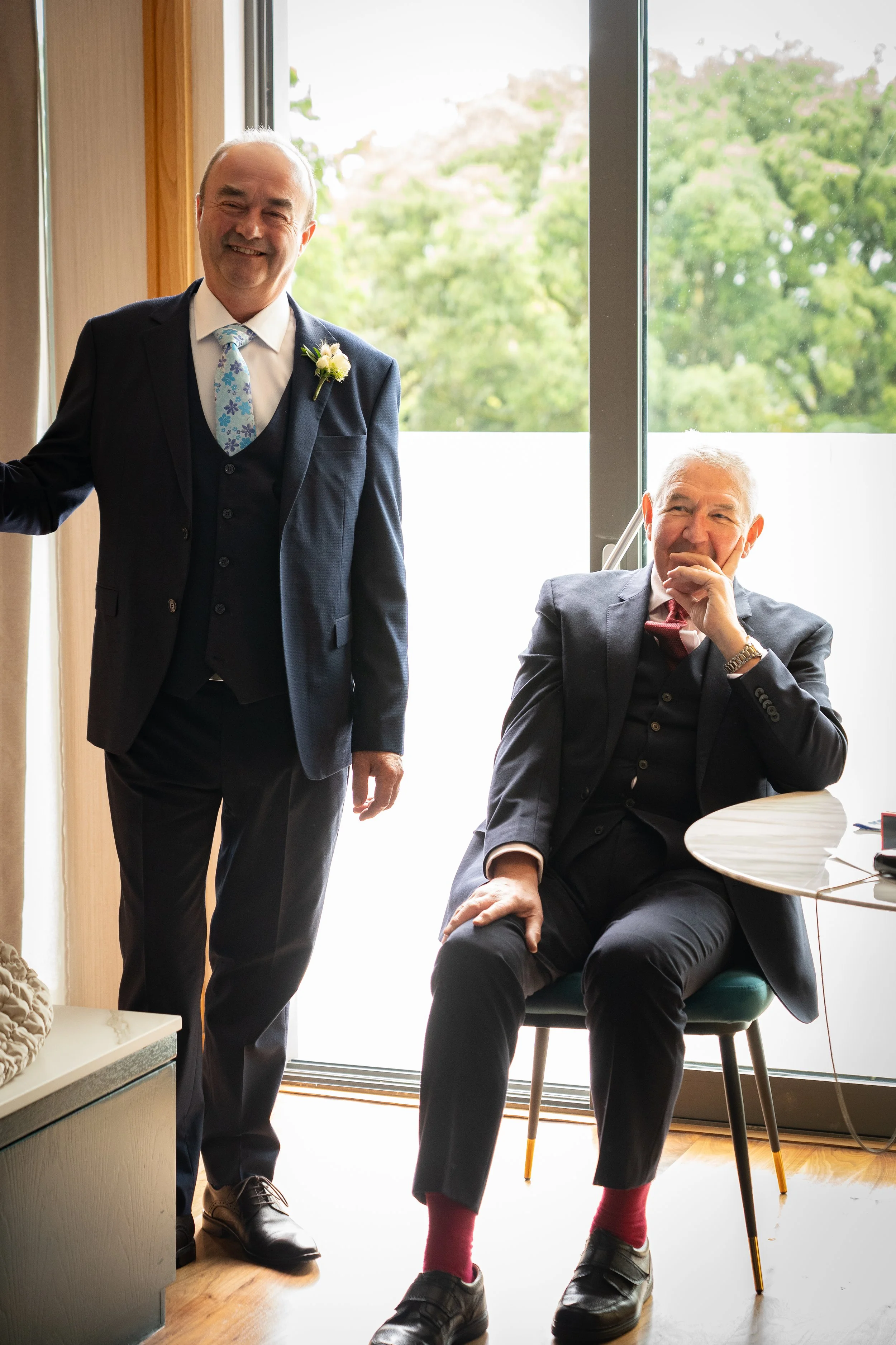 Two men in formal suits, one standing and smiling, the other sitting and laughing, inside a room with a large window showing green trees outside.