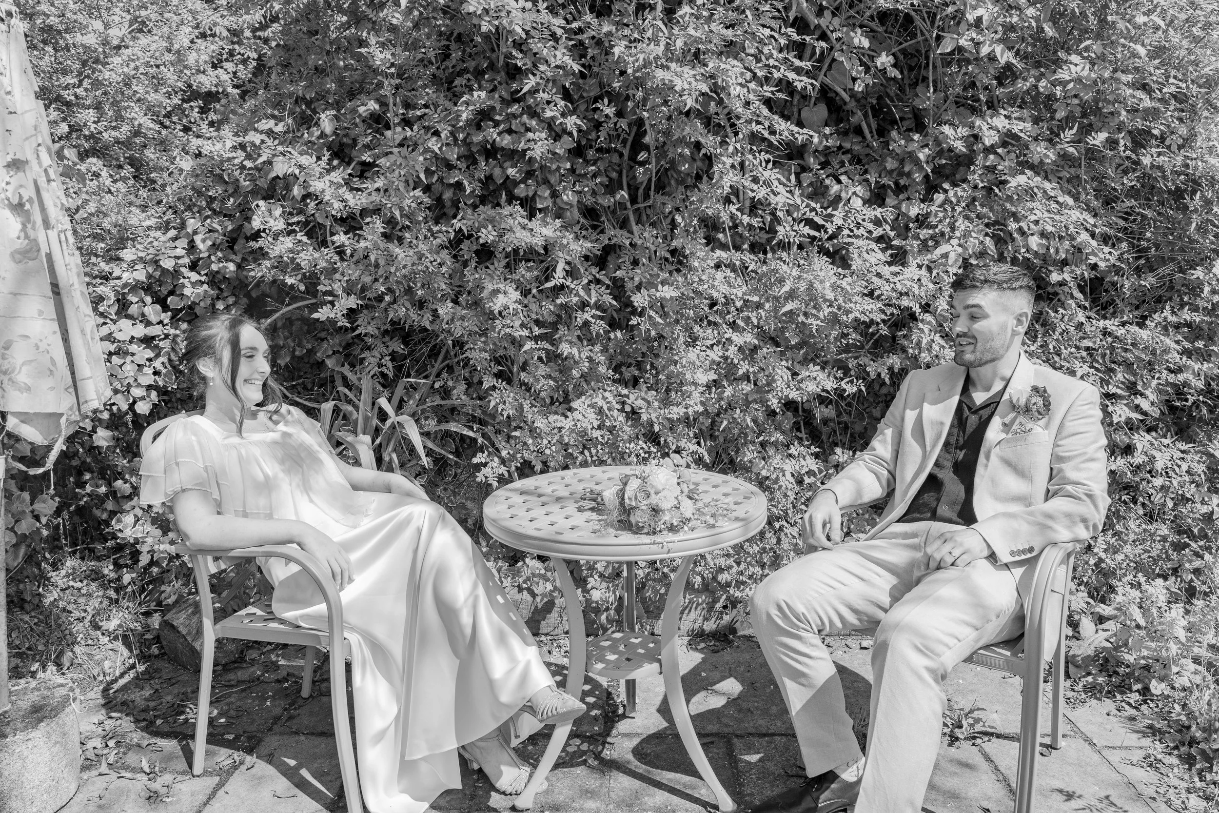 A black and white photo of a bride and groom sitting outdoors, smiling at each other. The bride is wearing a long dress, and the groom is in a light-colored suit. There is a small round table with a flower bouquet between them, and they are in front 