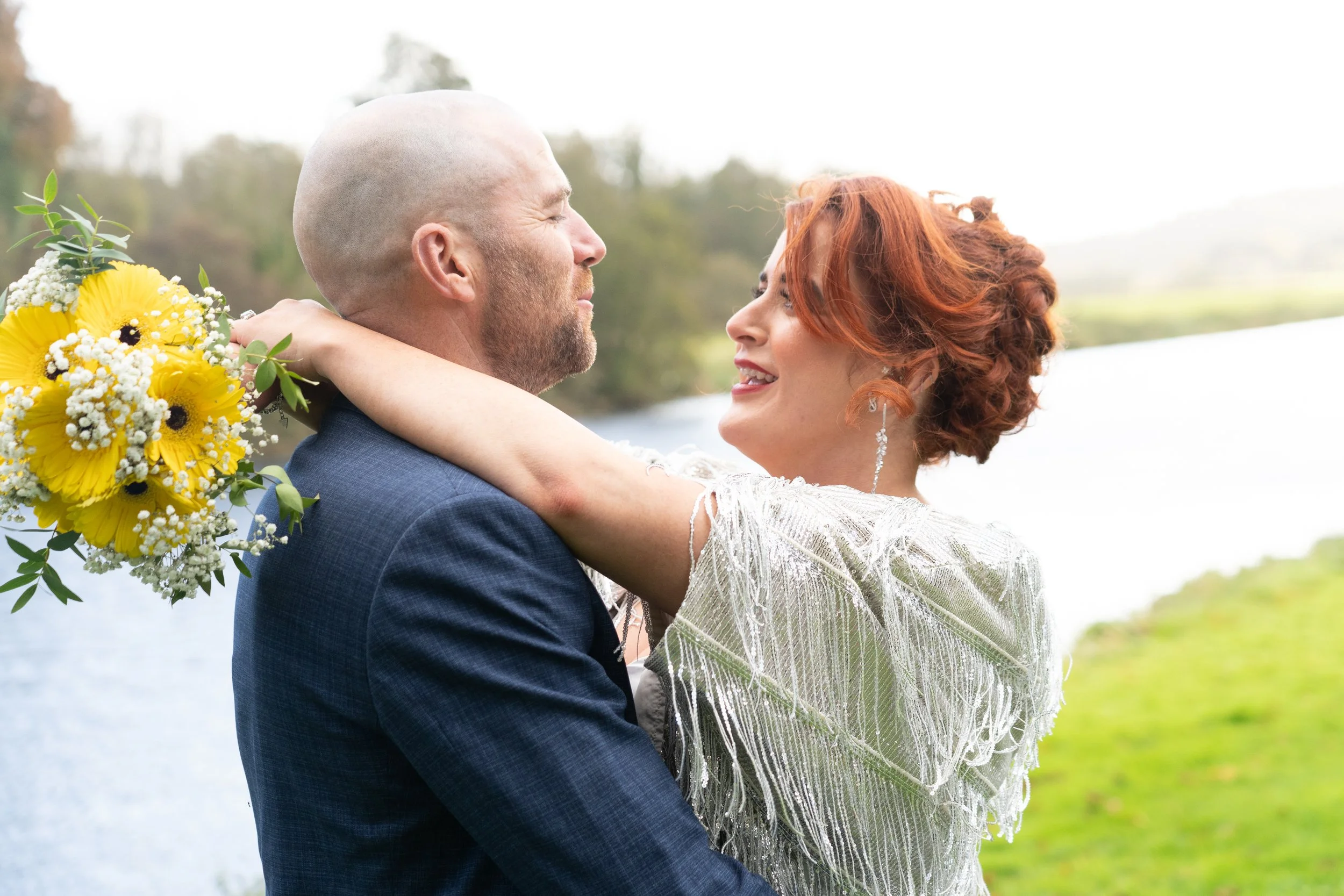 A couple dressed in wedding attire embracing outdoors near a body of water, with the woman holding a bouquet of yellow and white flowers.