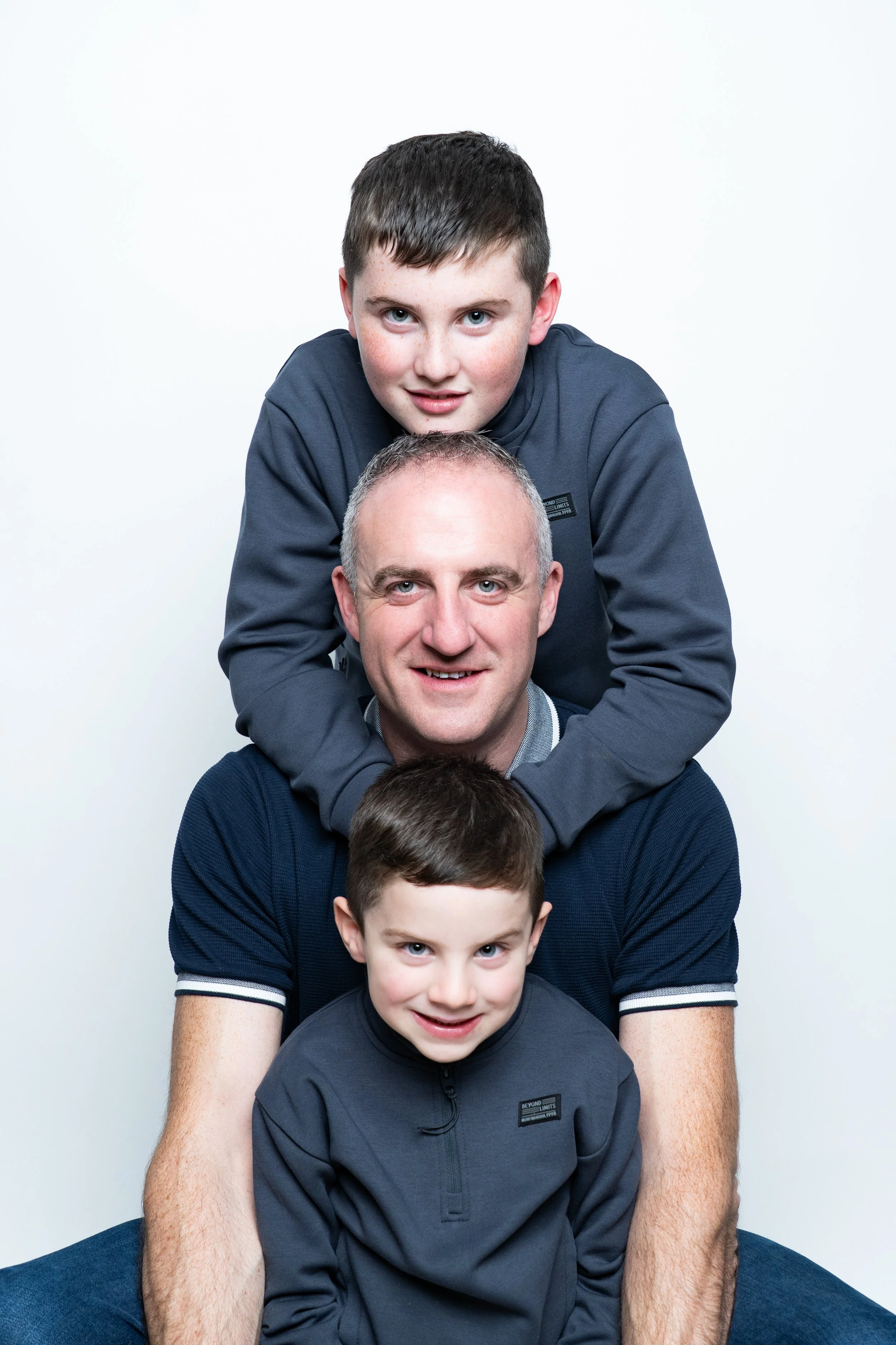 A family of four with two boys and a father, all smiling, posing together against a plain white background. The older boy is lying on top of the father, the younger boy is sitting in front of him, and the father is in the middle.