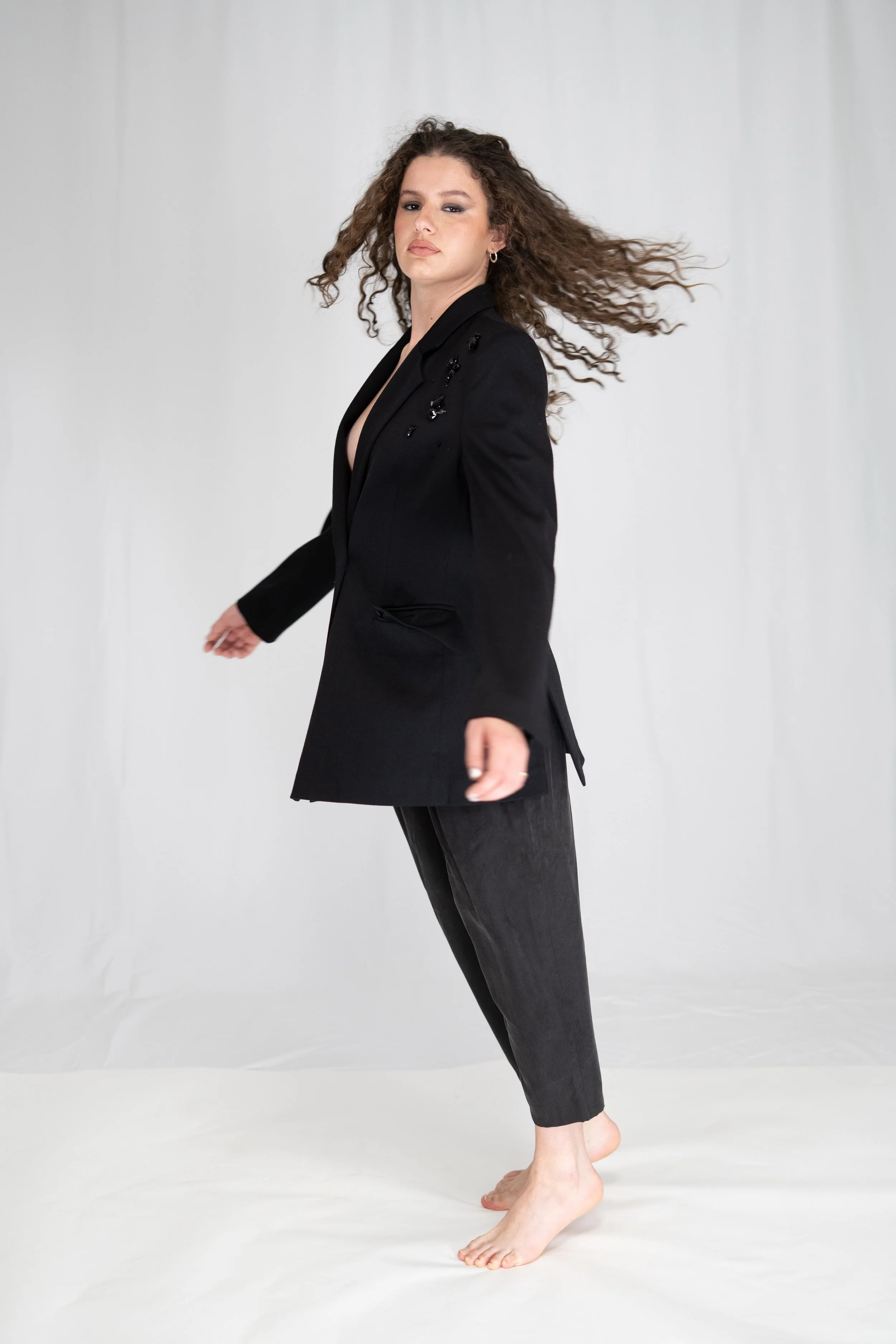 A woman with curly hair wearing a black blazer and dark pants standing barefoot against a plain white background.