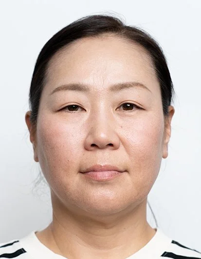 Portrait of a woman with black hair, wearing a white top with black stripes, looking directly at the camera against a plain light background.