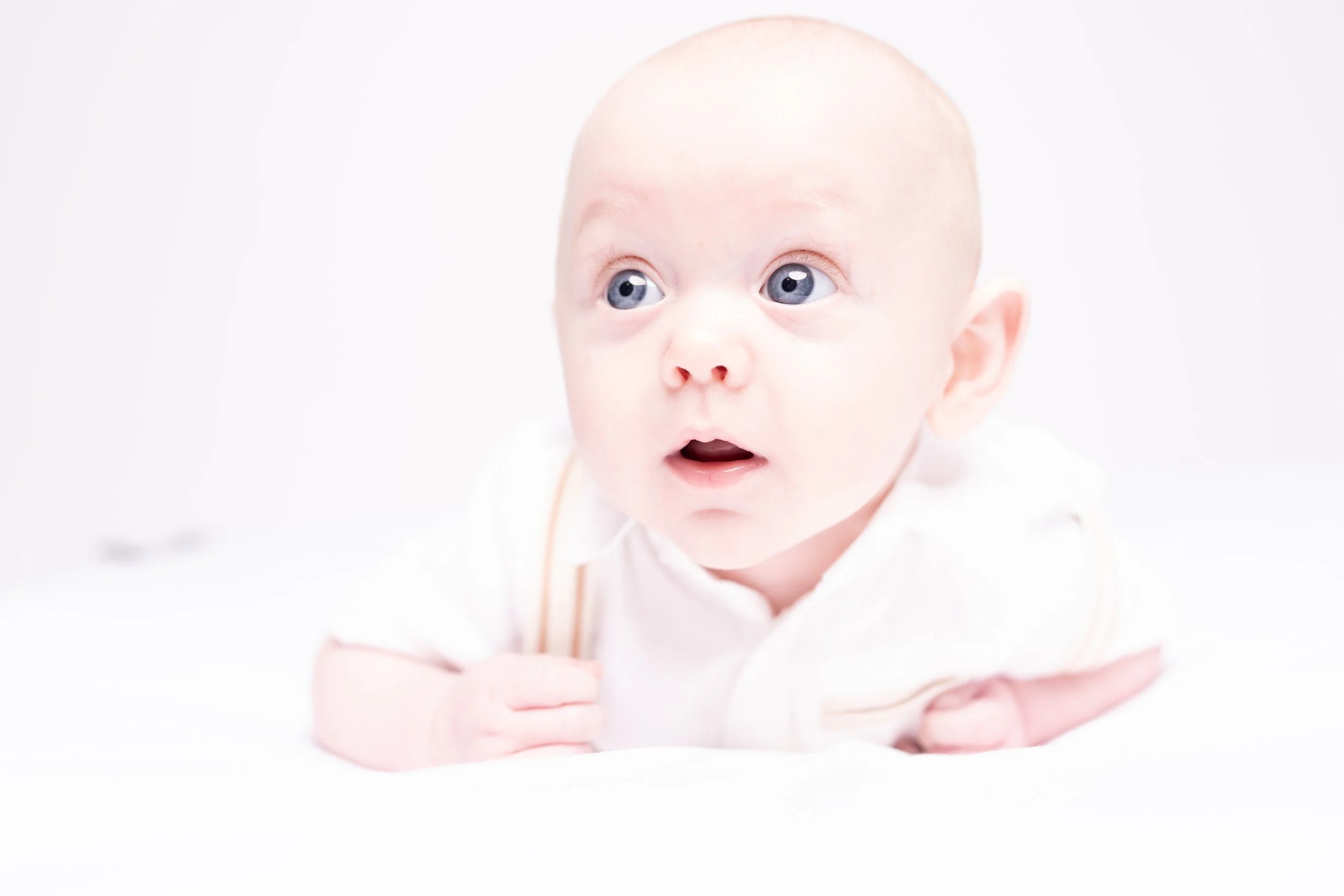 A close-up photo of a baby with blue eyes, lying on their stomach and looking upward. The baby has a surprised expression, is wearing a white outfit, and has a light background.