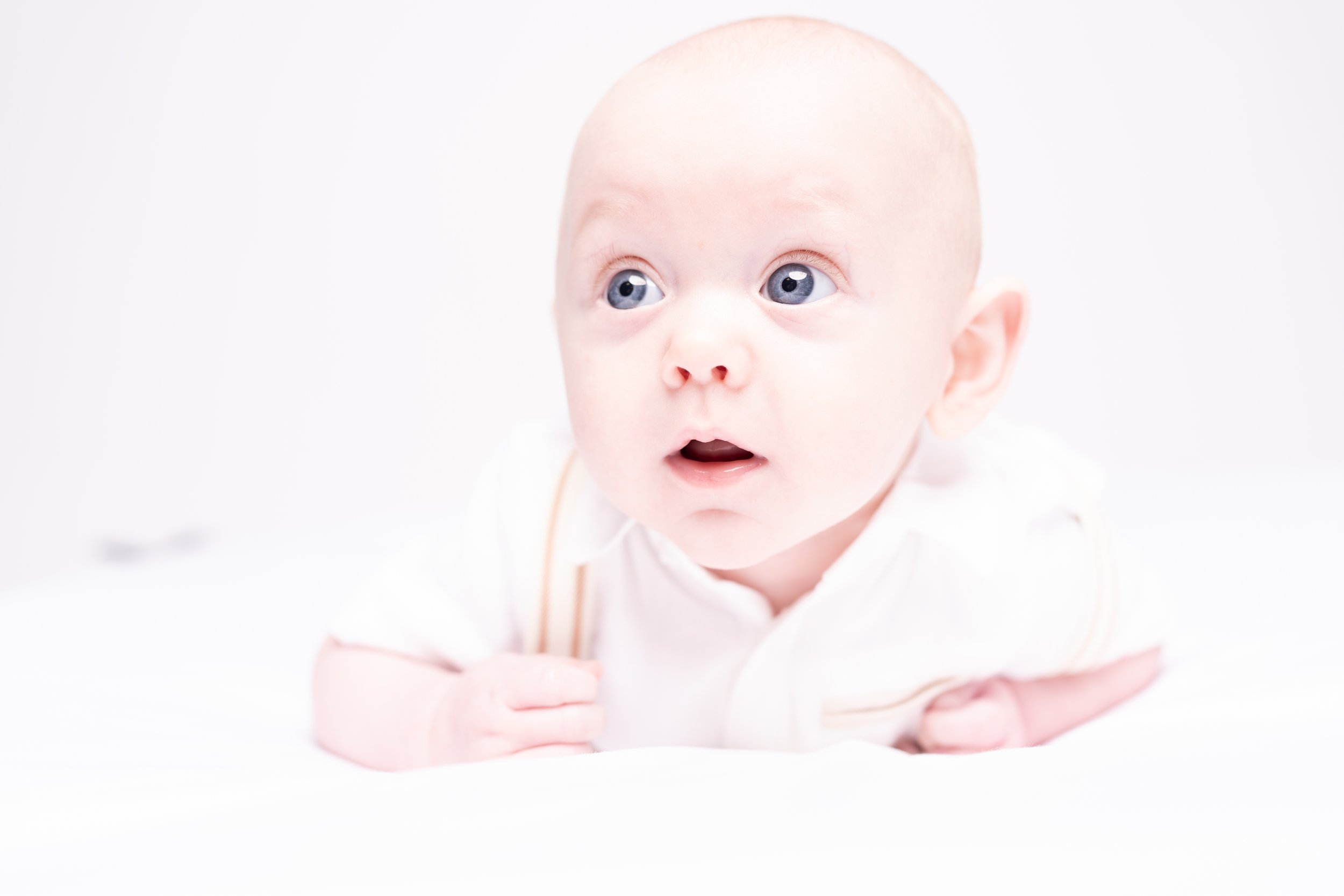 A close-up photo of a baby with blue eyes, lying on their stomach and looking upward. The baby has a surprised expression, is wearing a white outfit, and has a light background.