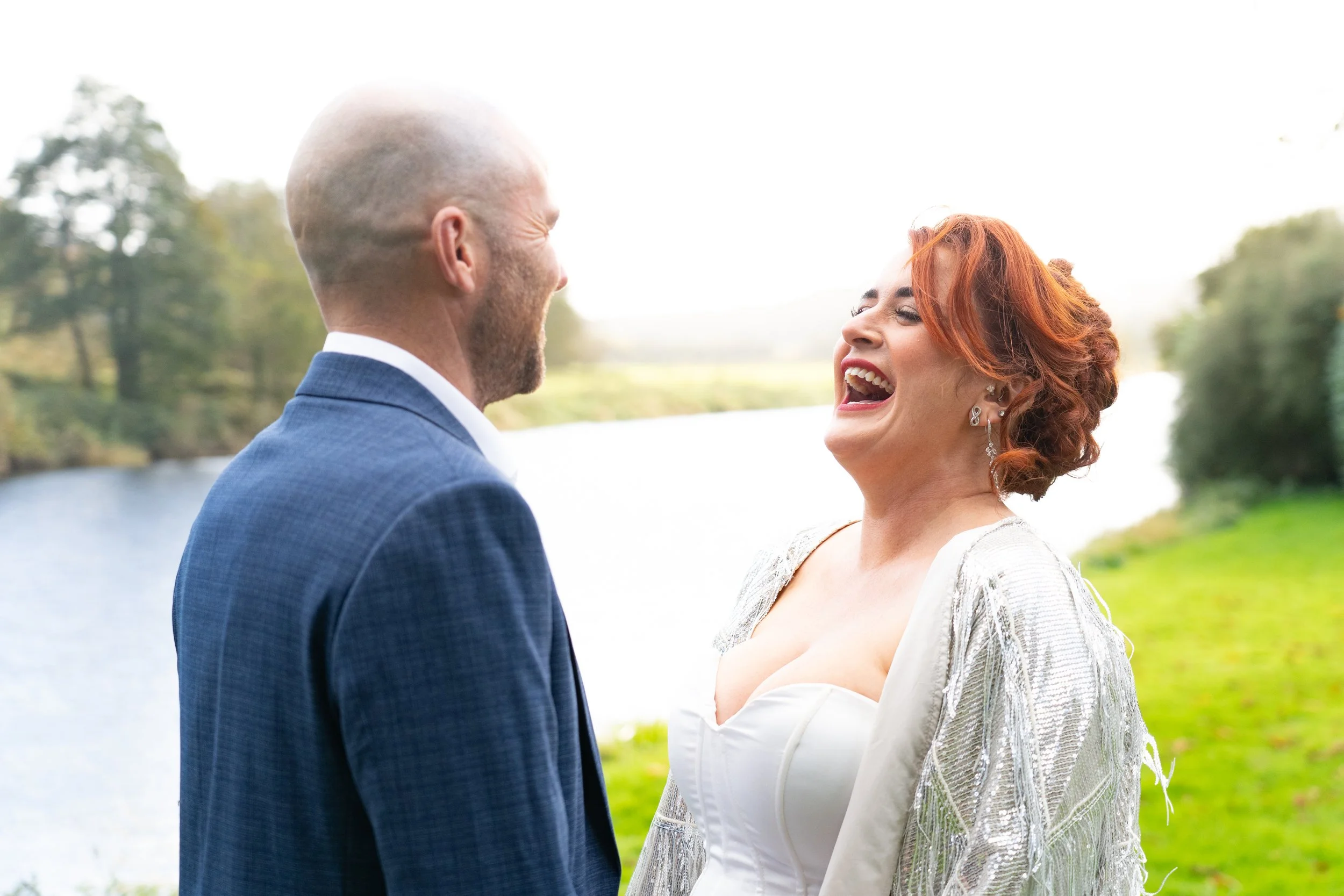 A happy couple laughing outdoors near a river, with trees in the background and bright lighting