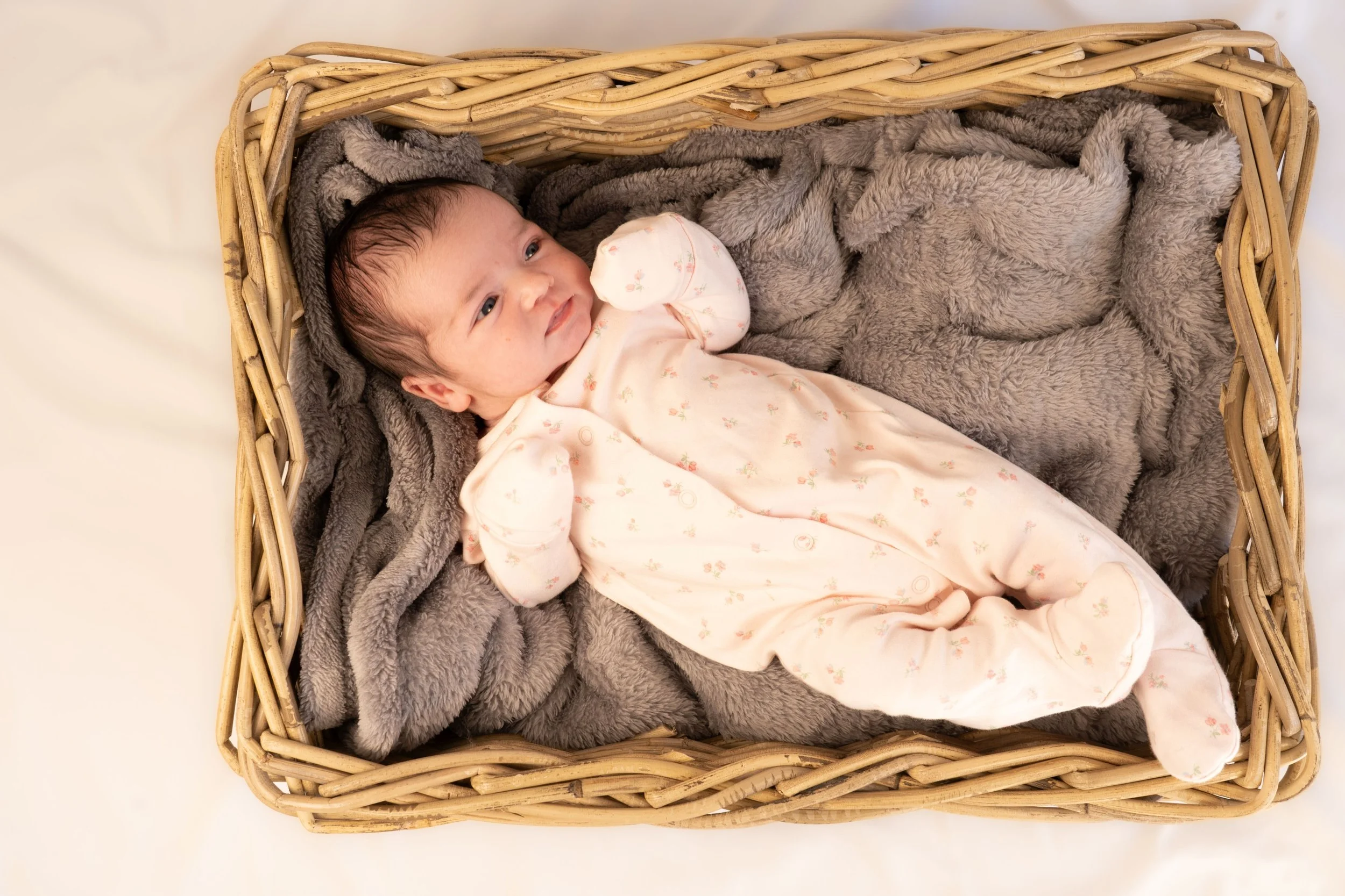 A baby lying in a wicker basket on gray blankets, wearing a pink and white floral sleeper.