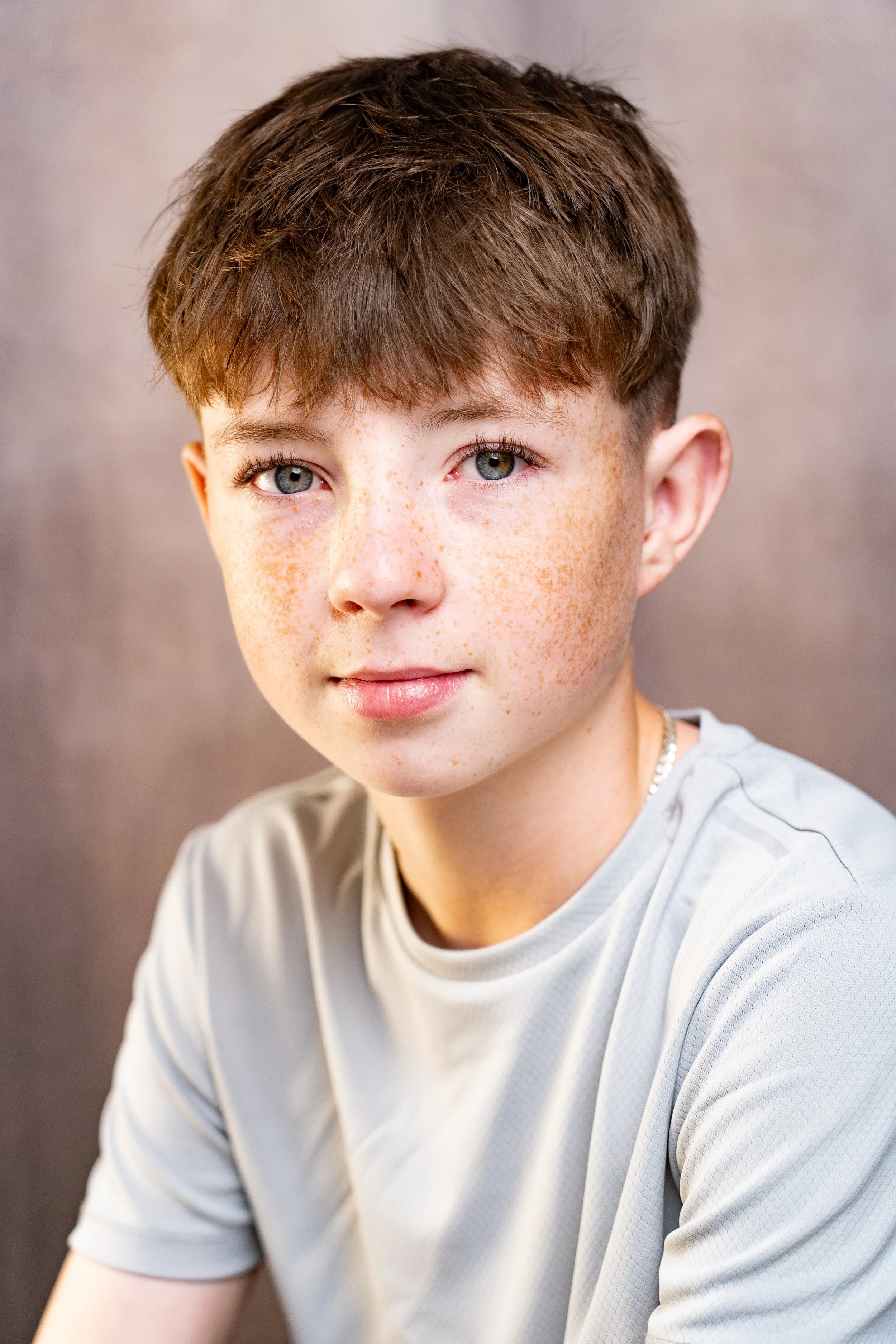 A close-up portrait of a young boy with brown hair, blue eyes, and freckles, wearing a light gray shirt, against a neutral background.
