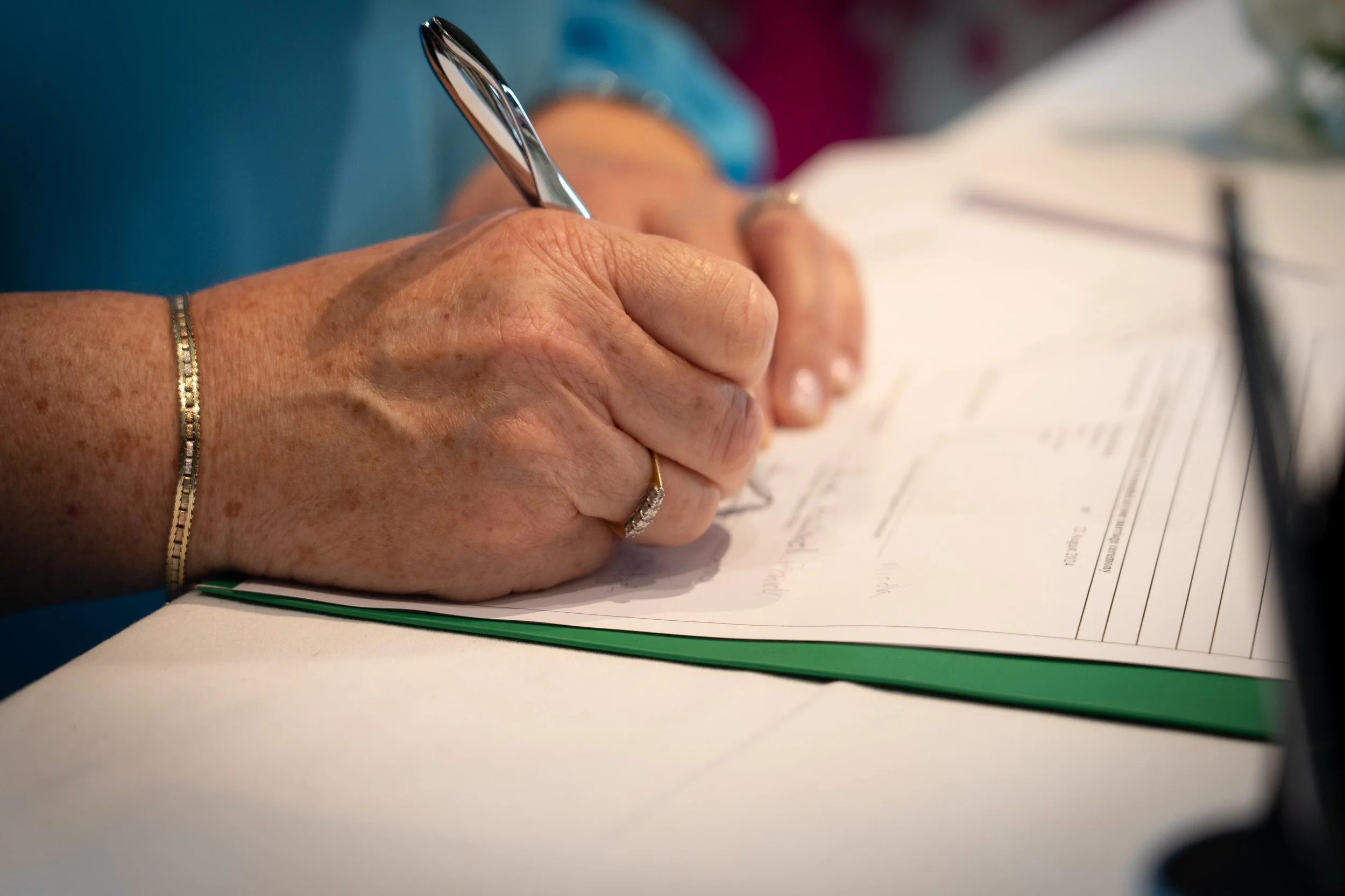 Close-up of an elderly person's hand signing a document with a pen, wearing a gold ring and a delicate bracelet.