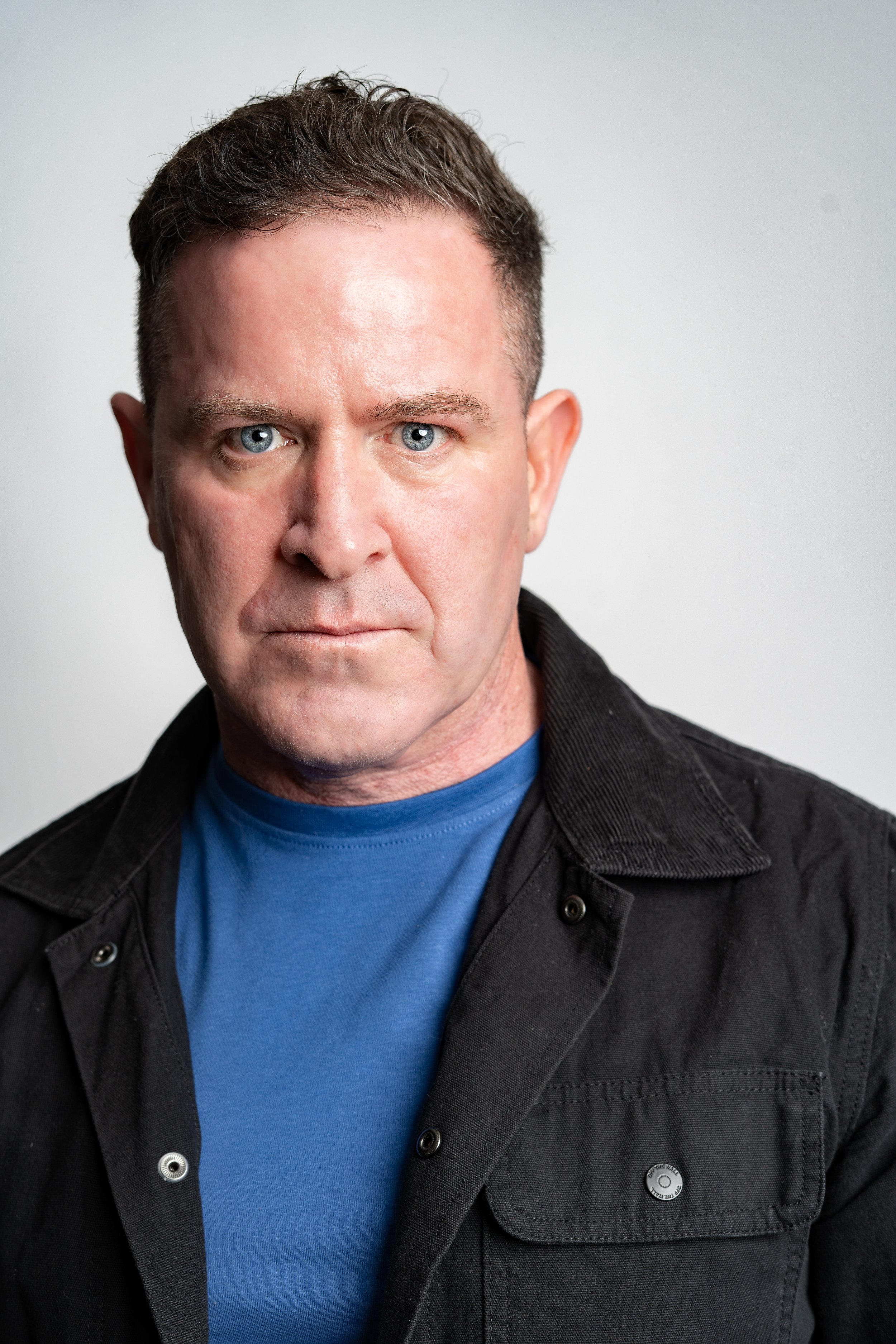 A man with short brown hair and blue eyes wearing a black jacket over a blue t-shirt, looking directly at the camera against a plain light gray background.