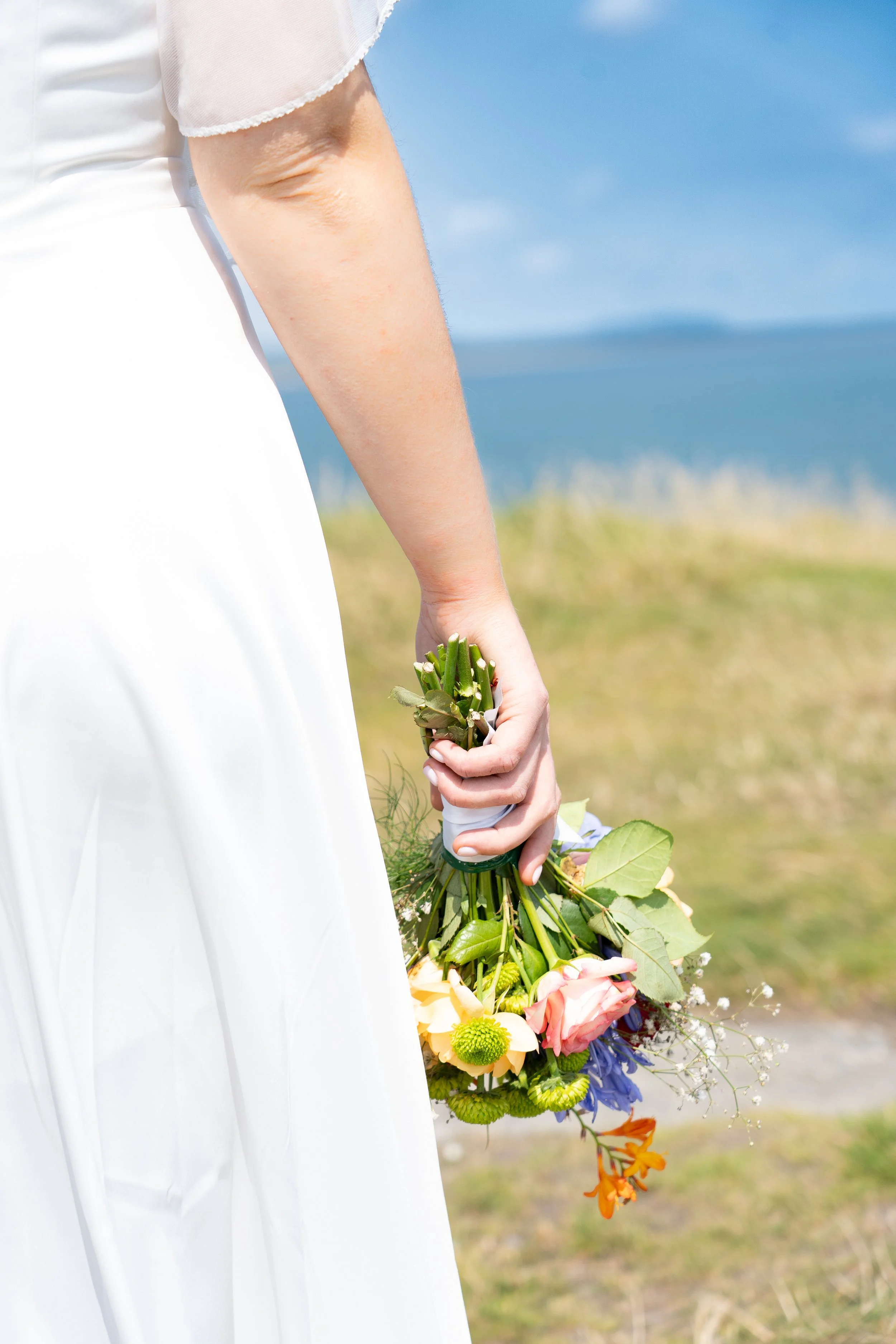 A person in a white dress holding a small bouquet of various colorful flowers, standing outdoors near a body of water and grassy landscape.