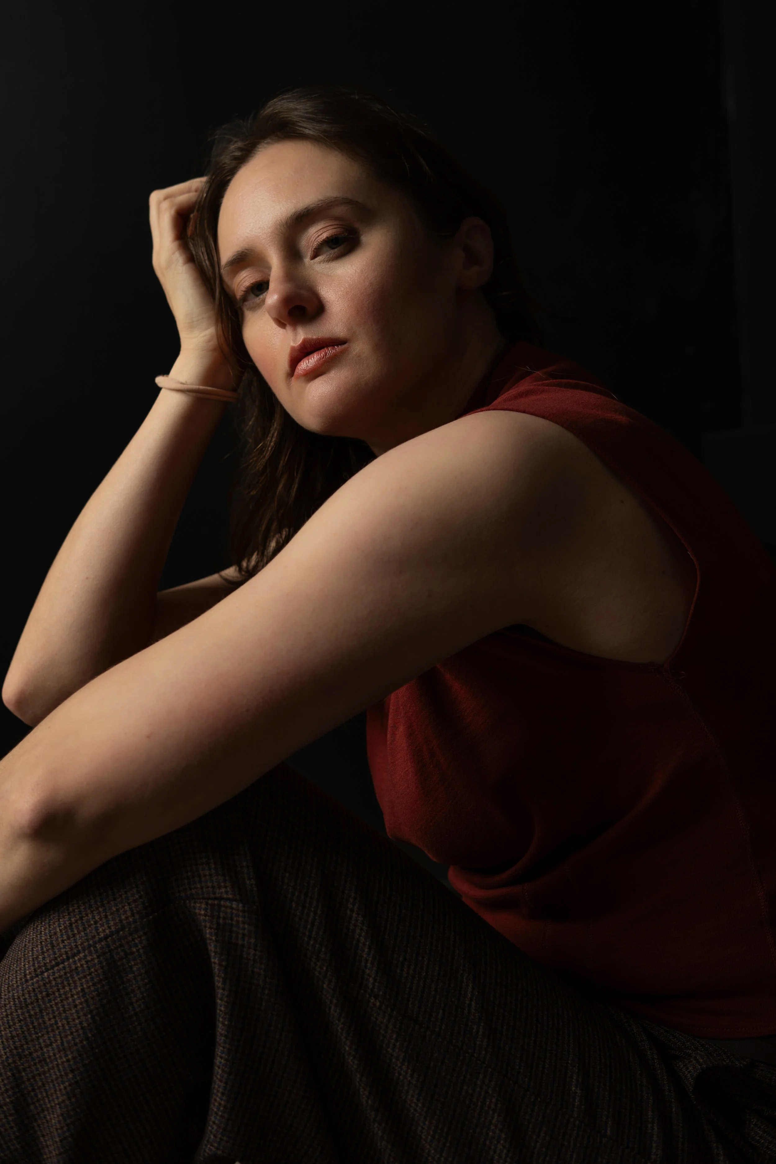 A woman with shoulder-length brown hair, wearing a sleeveless red top, sitting against a dark background, looking at the camera with a serious expression.