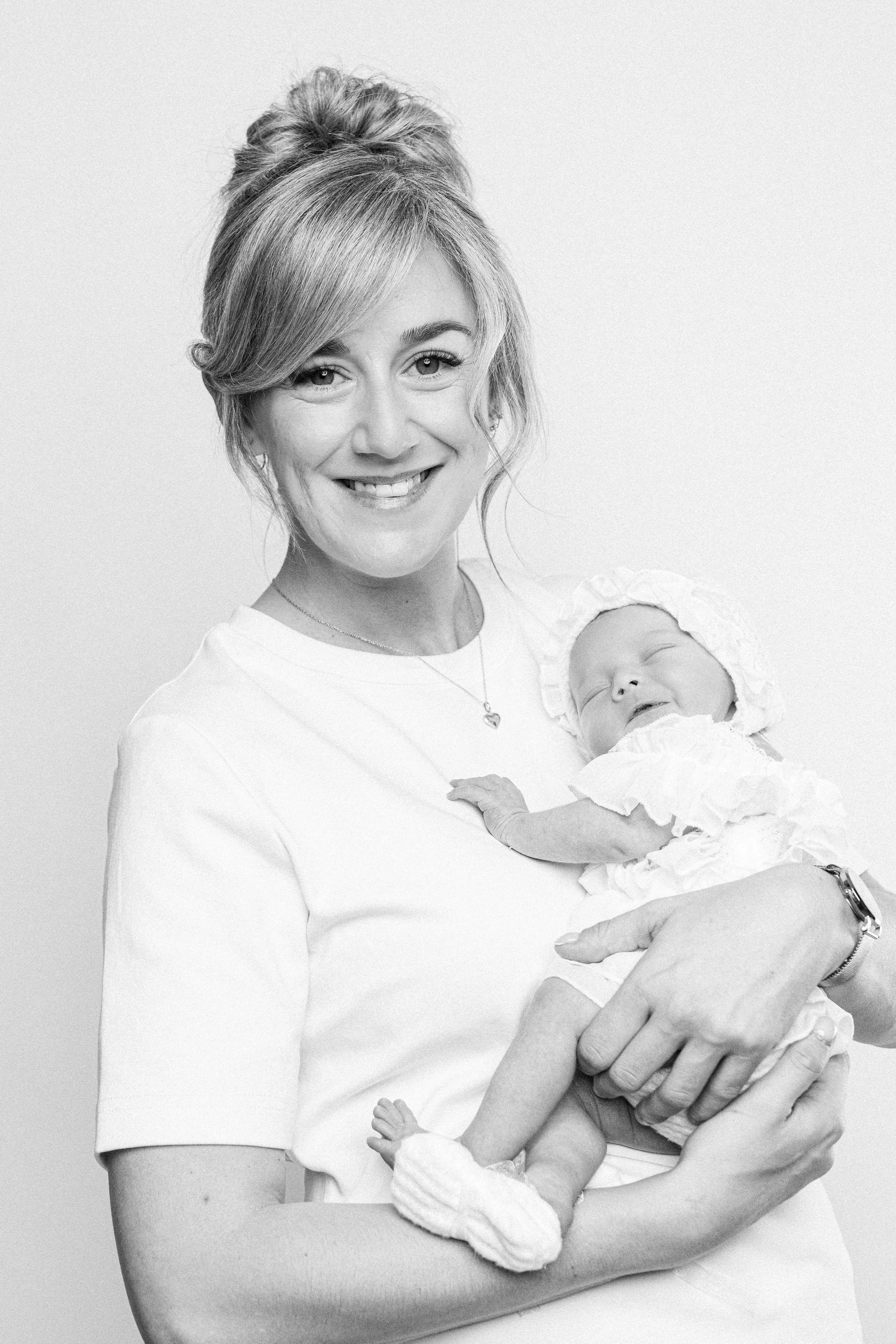 A woman smiling and holding a sleeping baby girl dressed in white.