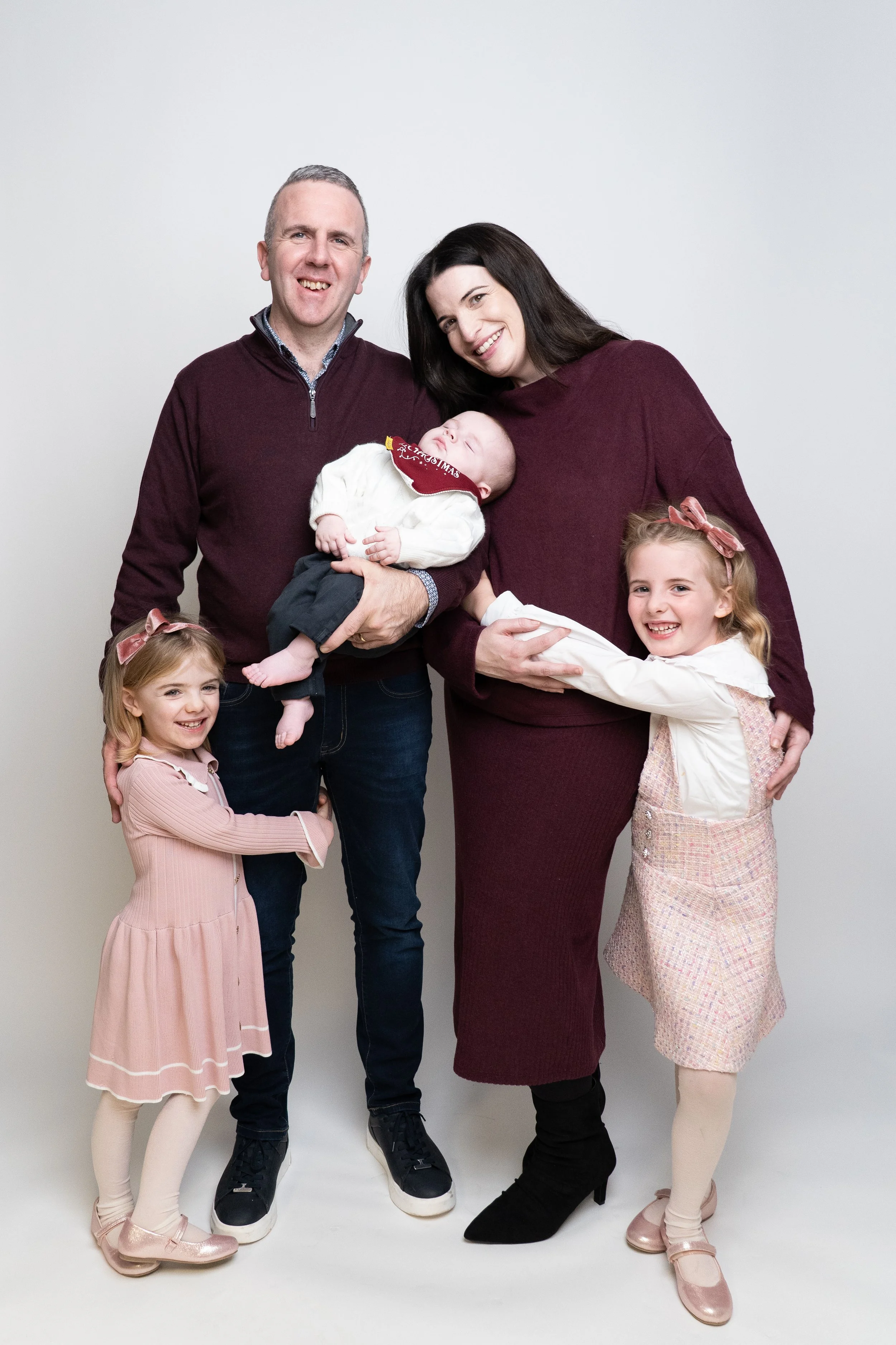 Family photo of two adults and three children posing together against a white backdrop, with two girls smiling and a baby being held by a man. The woman is holding the baby's legs, and the children are dressed in festive, pastel, and pink clothing.
