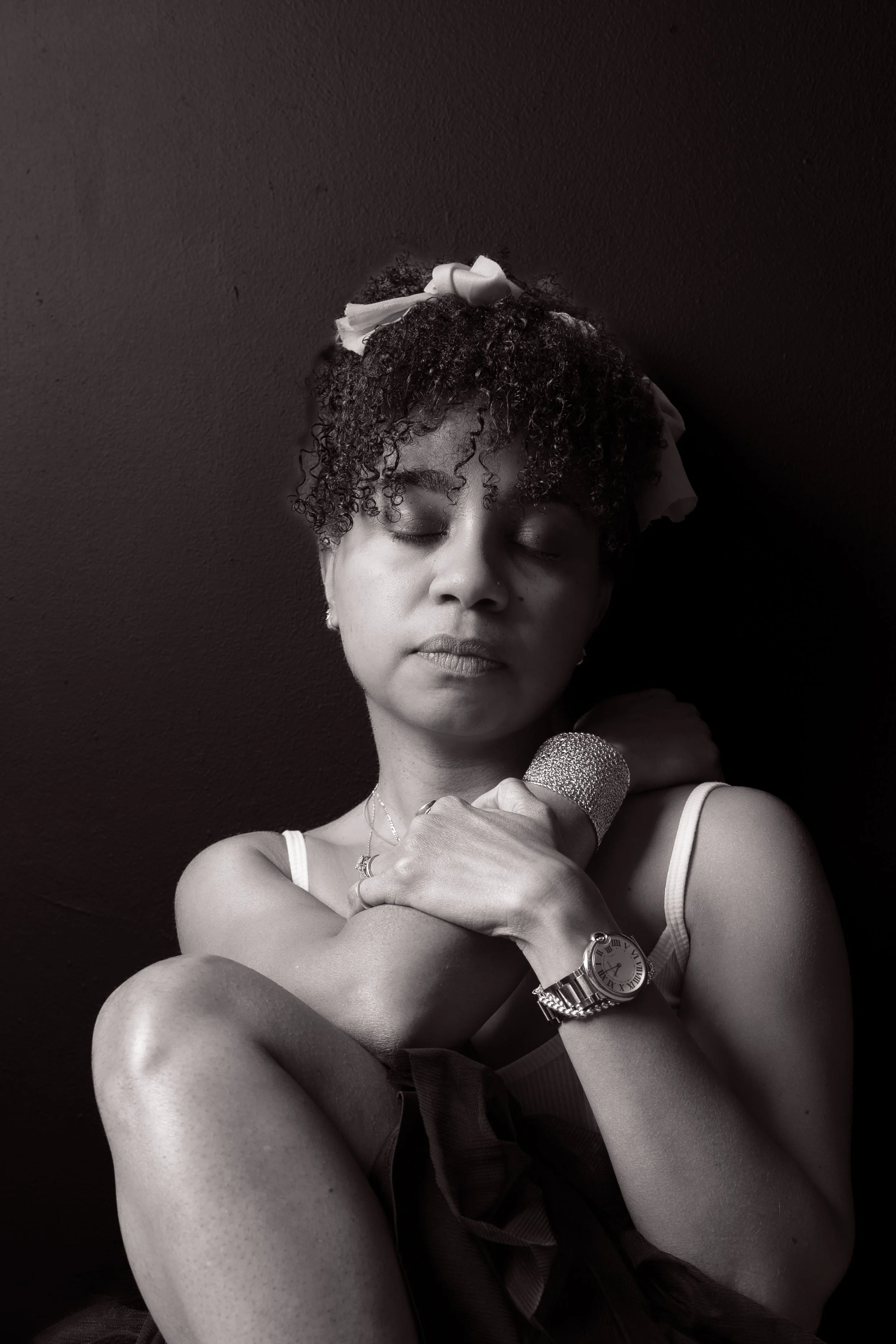 A woman with curly hair, a bow, and jewelry is holding a microphone with her eyes closed against a dark background.