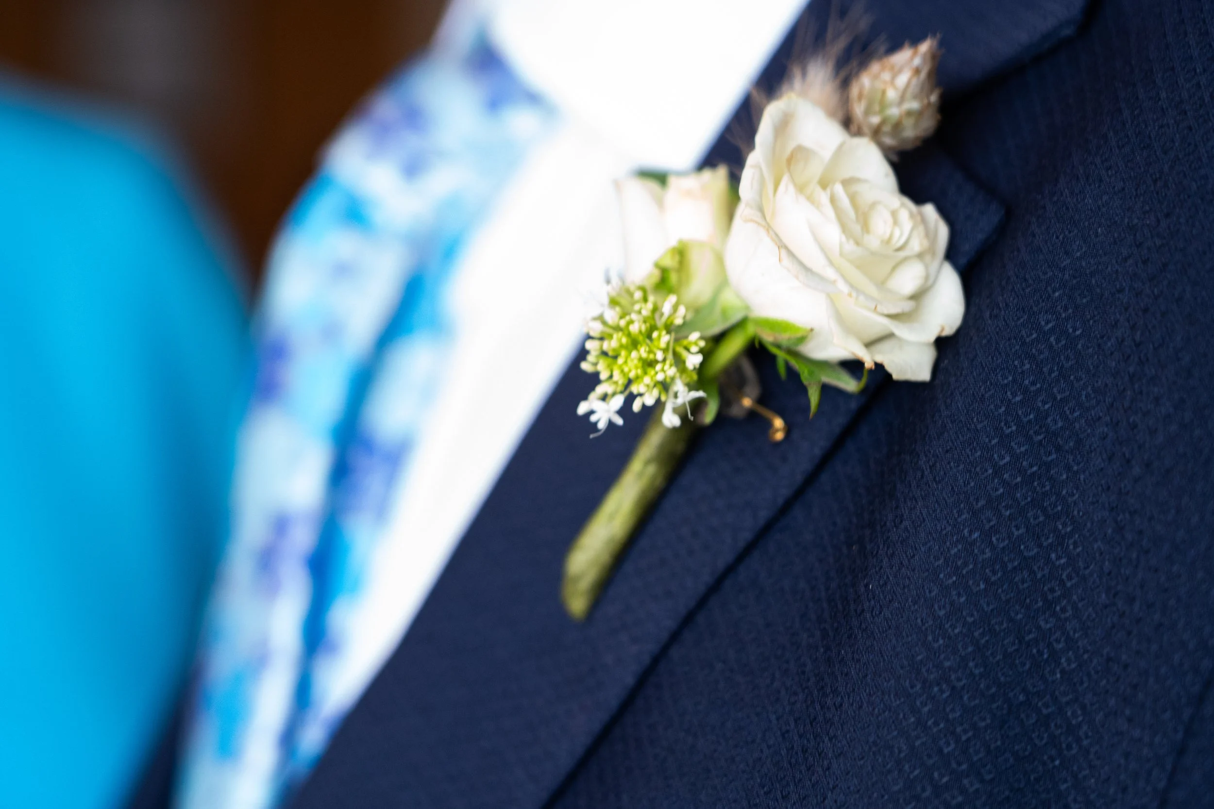 Close-up of a wedding boutonniere with white roses, small green flowers, and green foliage pinned to a navy blue suit.