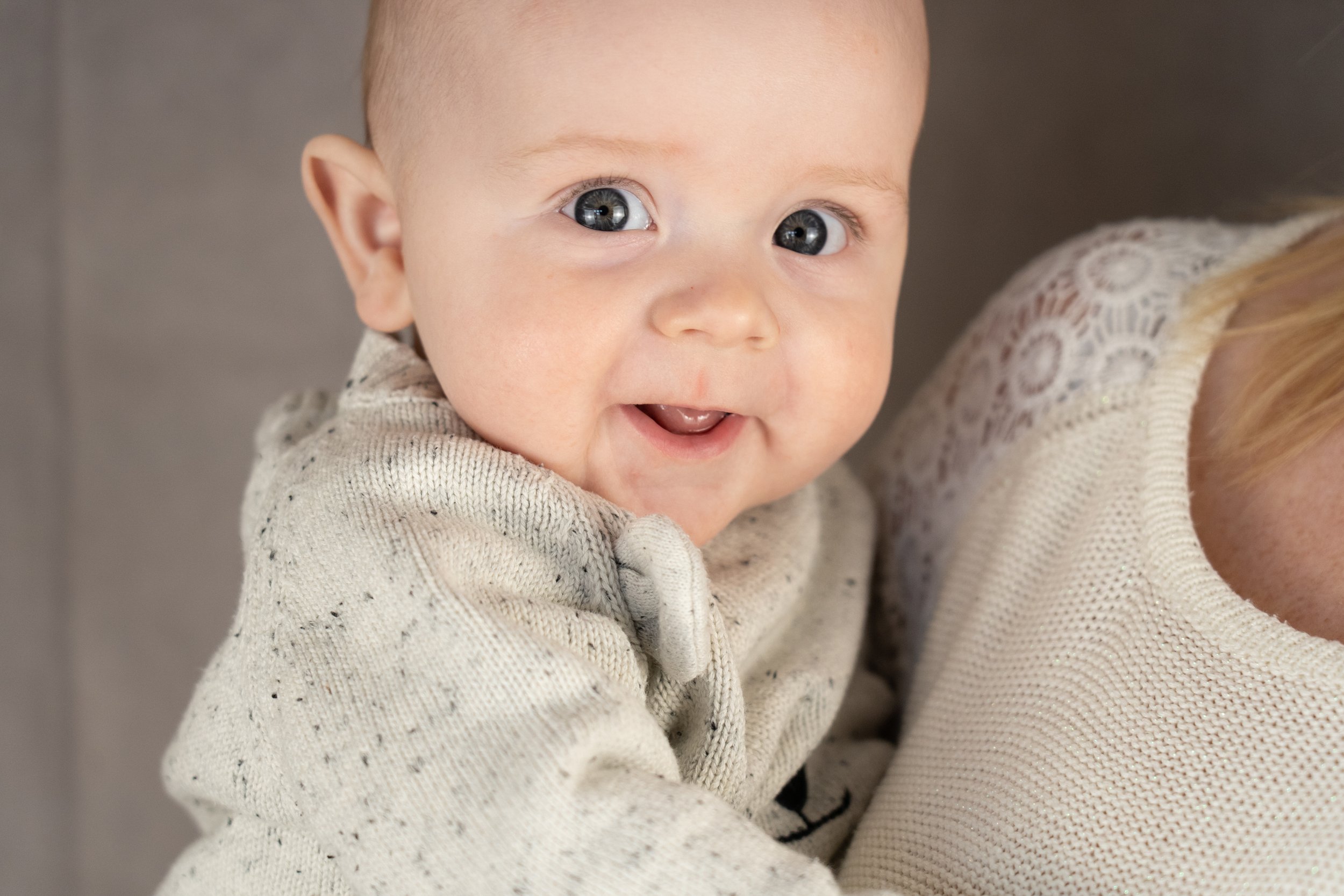 Close-up of a smiling baby with blue eyes, wearing a cream-colored sweater, being held by a person wearing a beige textured top.