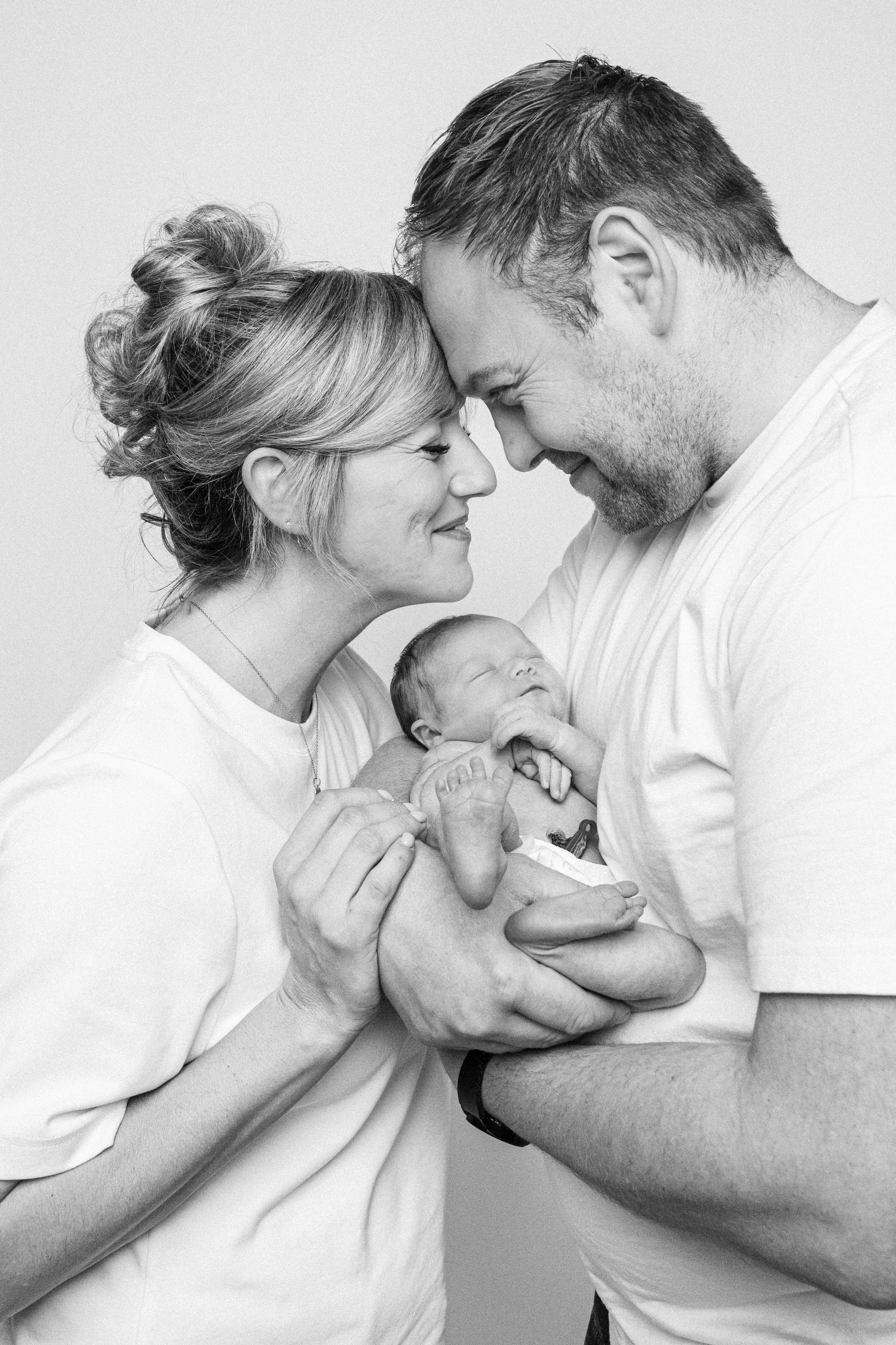 Black and white photograph of a family with a newborn. The mother and father are touching their foreheads together, smiling lovingly. The mother is holding the baby close to her chest, and the father is supporting the baby's body with his arms. The b