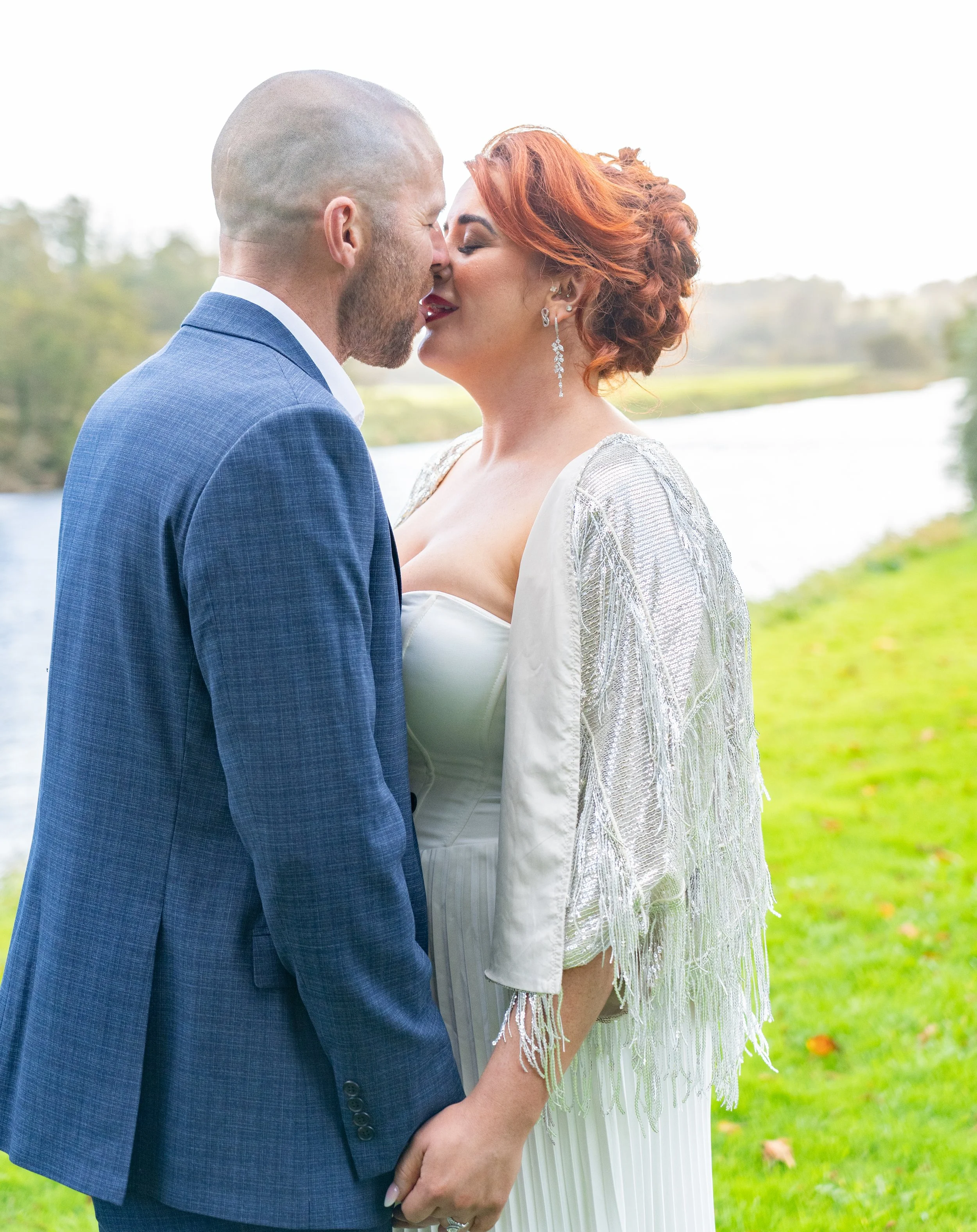 A couple dressed in wedding attire kissing outdoors near a river with green grass and trees in the background.