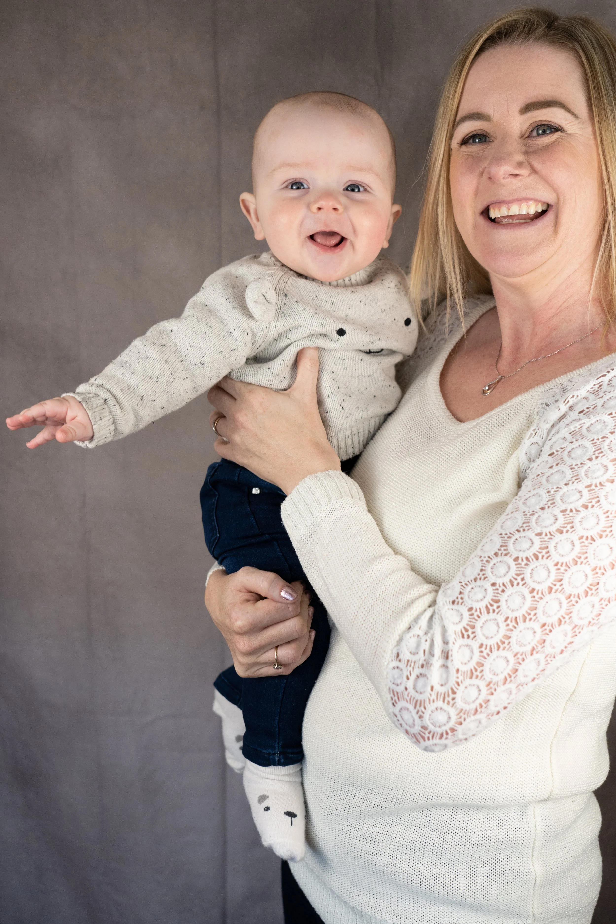 A woman holding a smiling baby with a gray background.