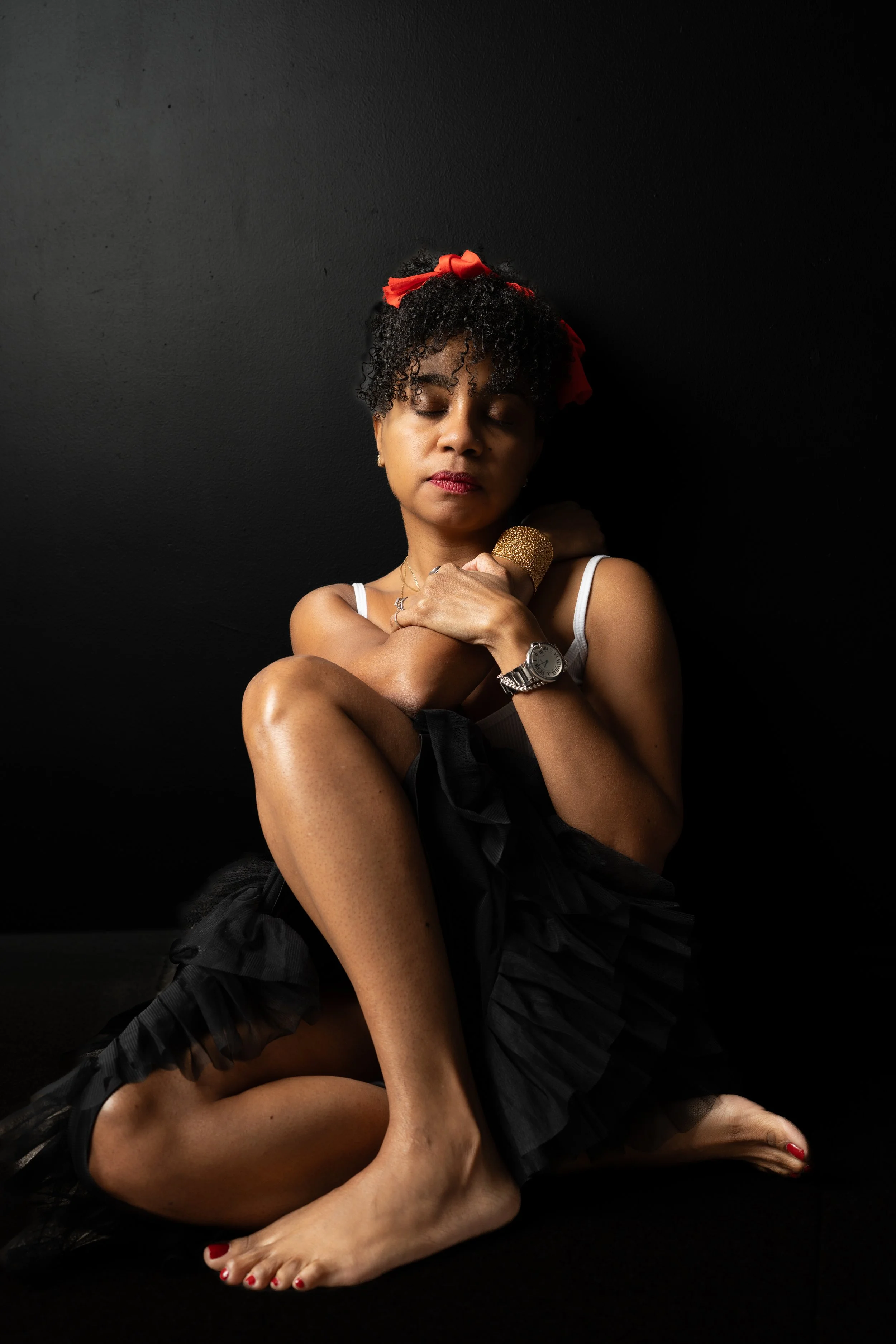 A woman with curly hair, a red headband, and red nail polish sitting against a black wall, hugging herself with her eyes closed.