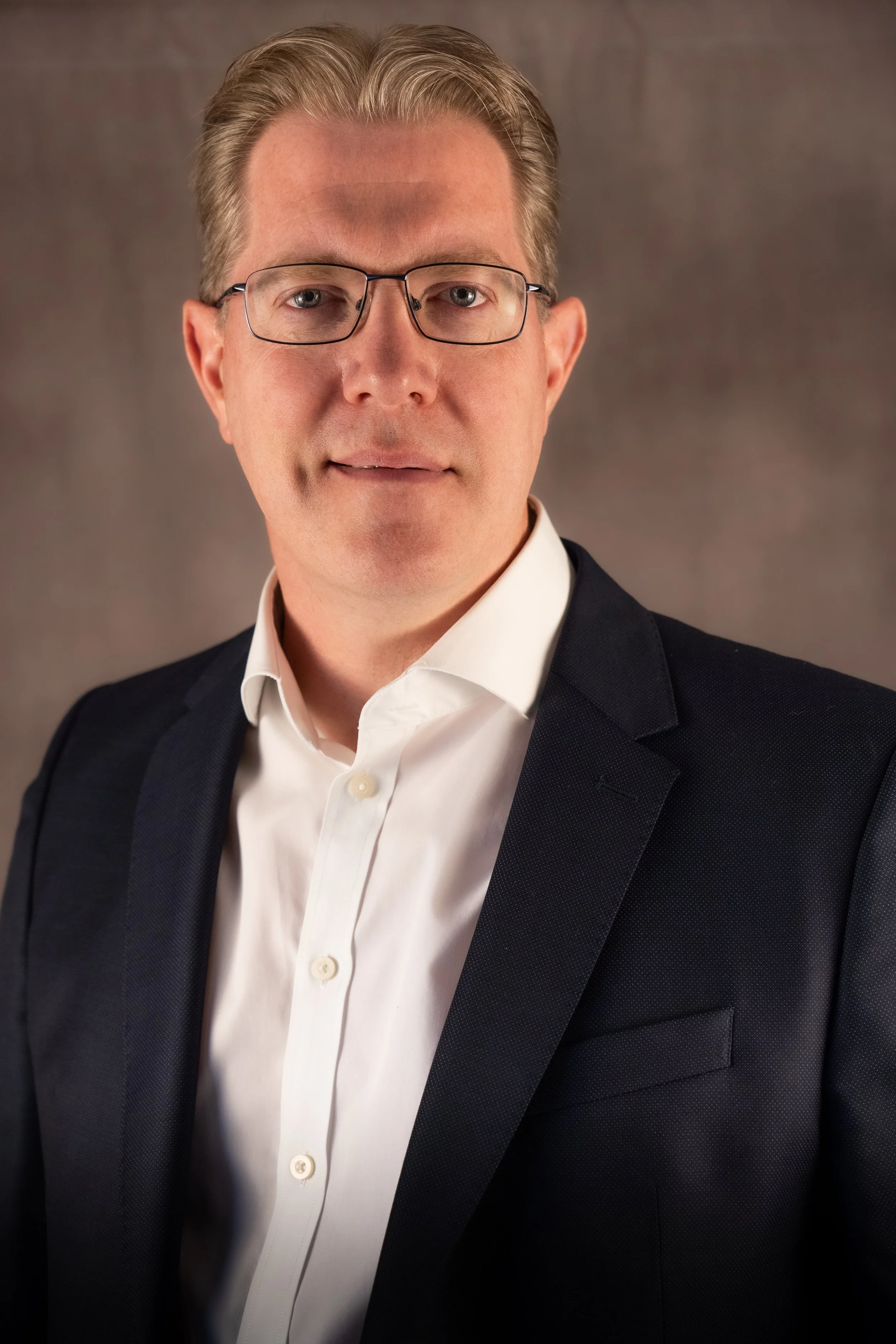 A portrait of a man with light hair, wearing glasses, a white shirt, and a dark blazer, against a brownish background.