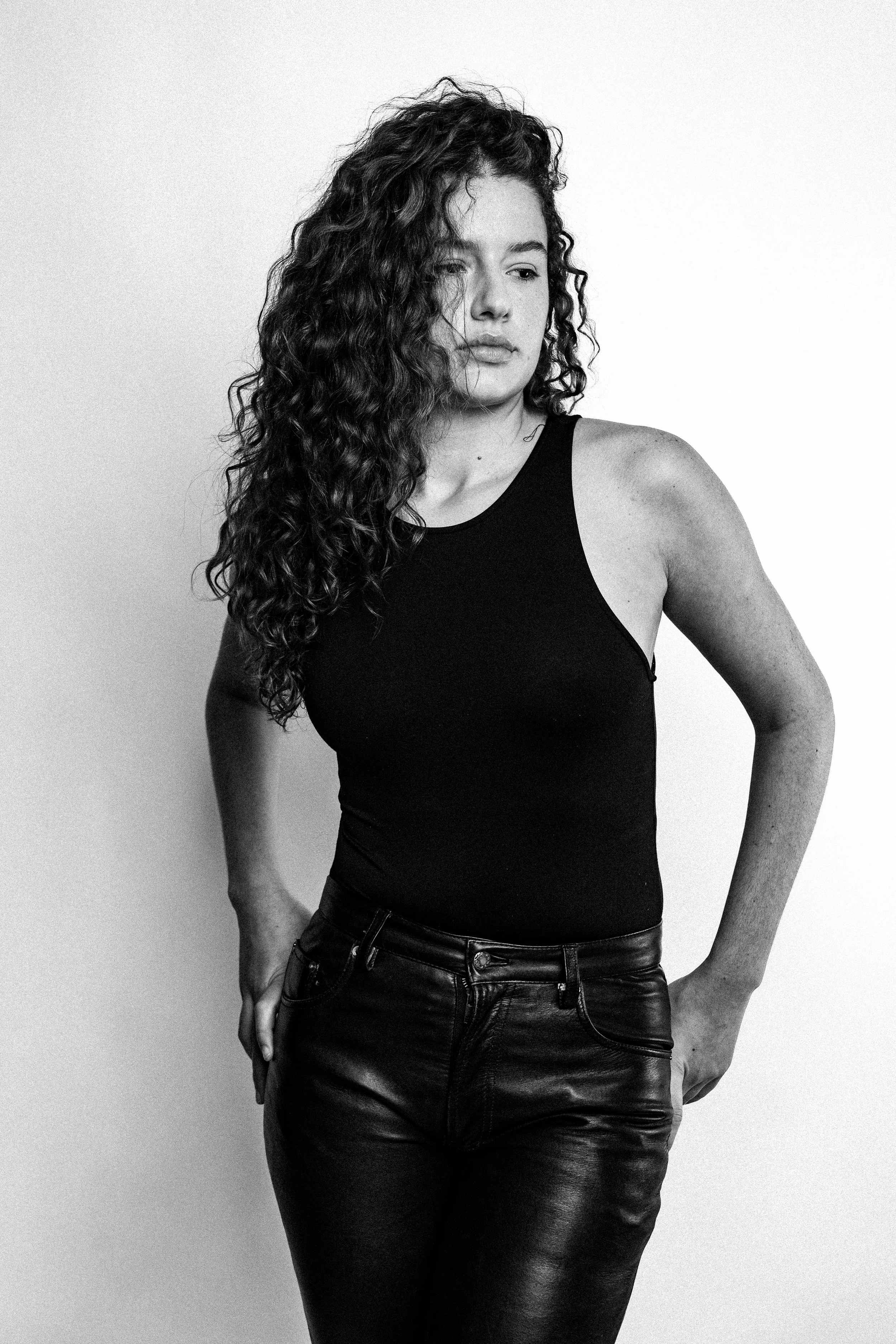 Black and white portrait of a woman with curly hair, wearing a sleeveless top and leather pants, standing against a plain background.