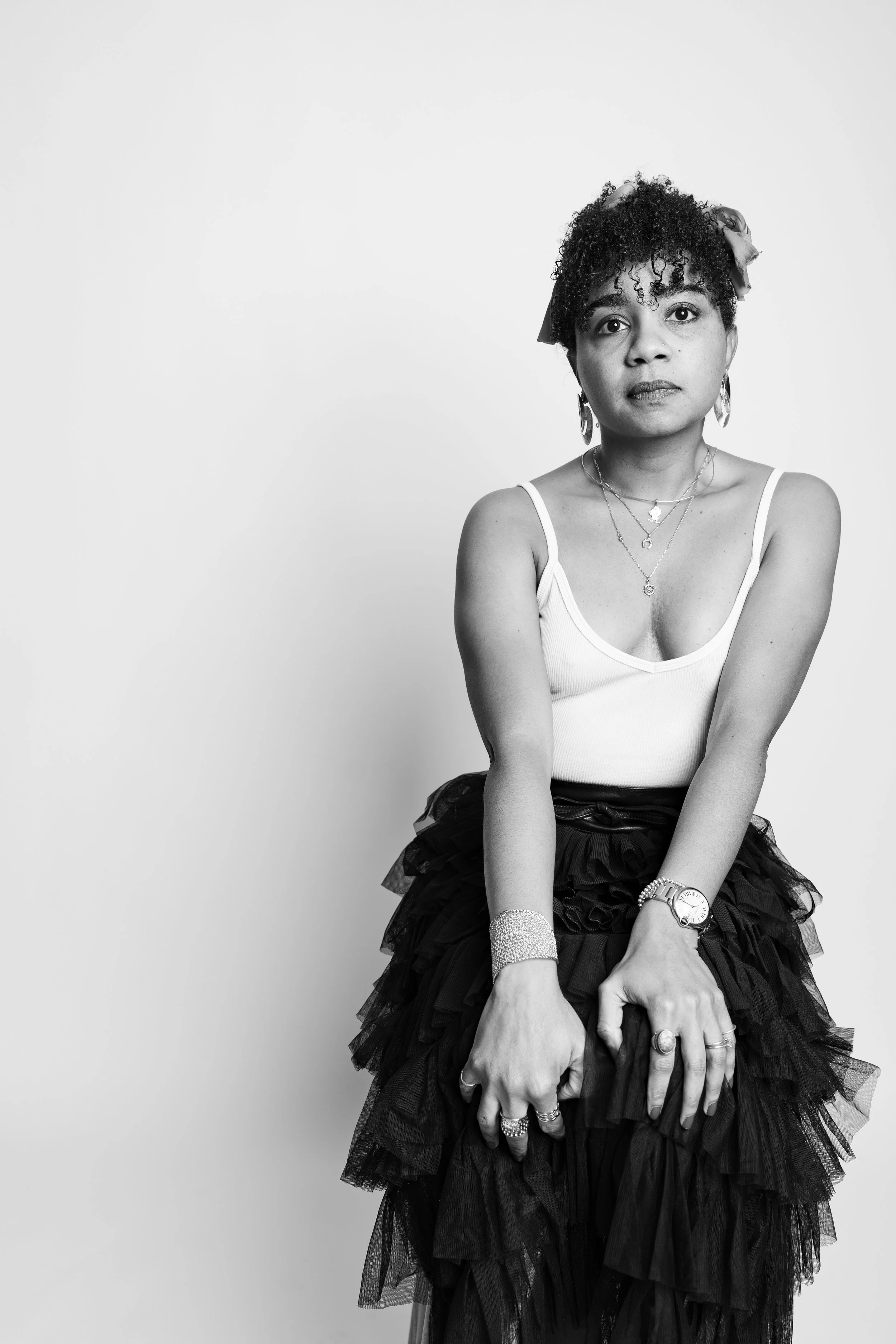 Black and white photo of a woman with short, curly hair, wearing earrings, layered necklaces, and a white tank top with thin straps, paired with a layered, ruffled black skirt. She has her hands clasped in front and wears multiple rings, a watch, and