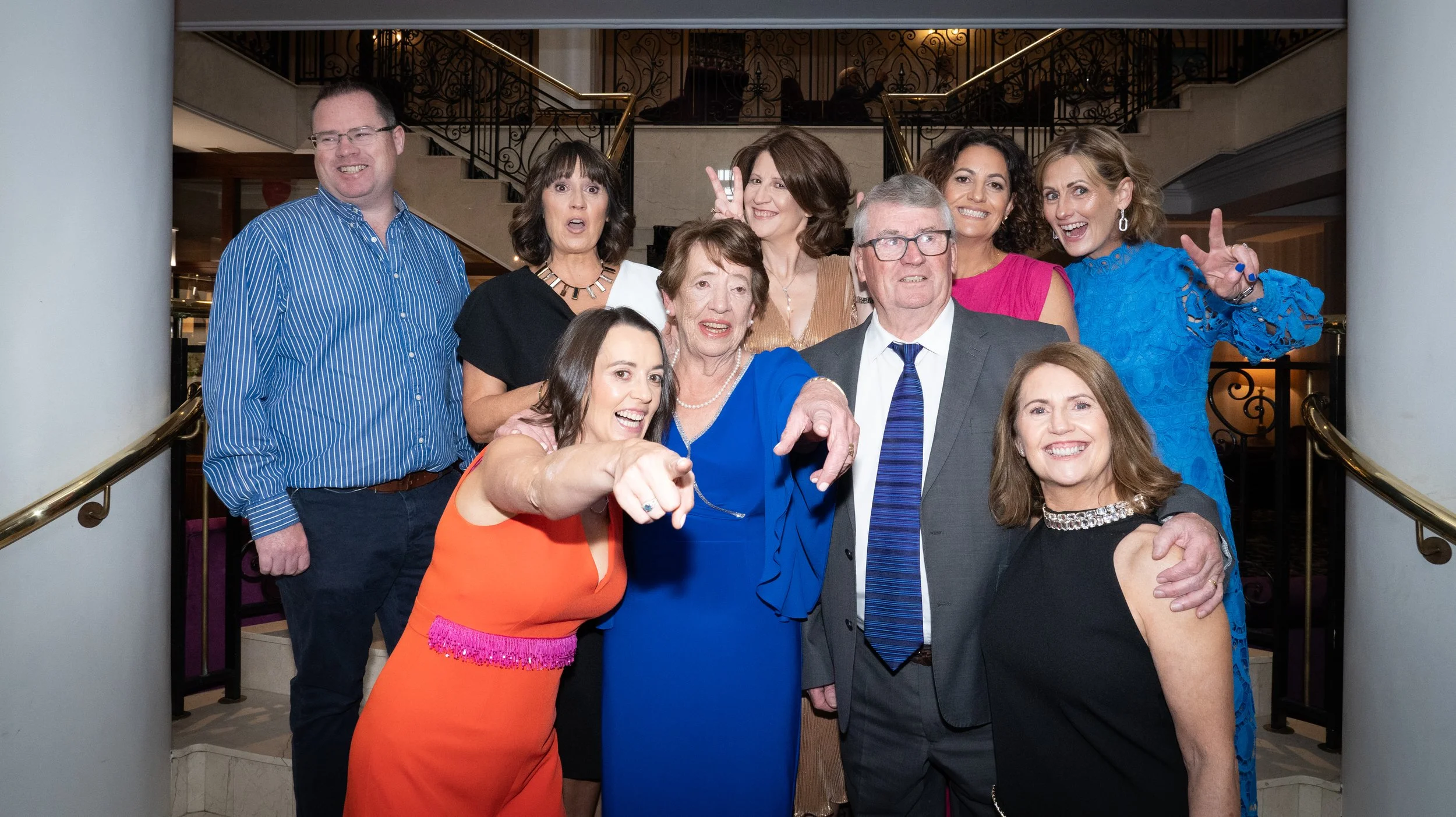 A group of ten people posing together at an indoor event, with some pointing and making peace signs, standing on a staircase with ornate metal railings in the background.