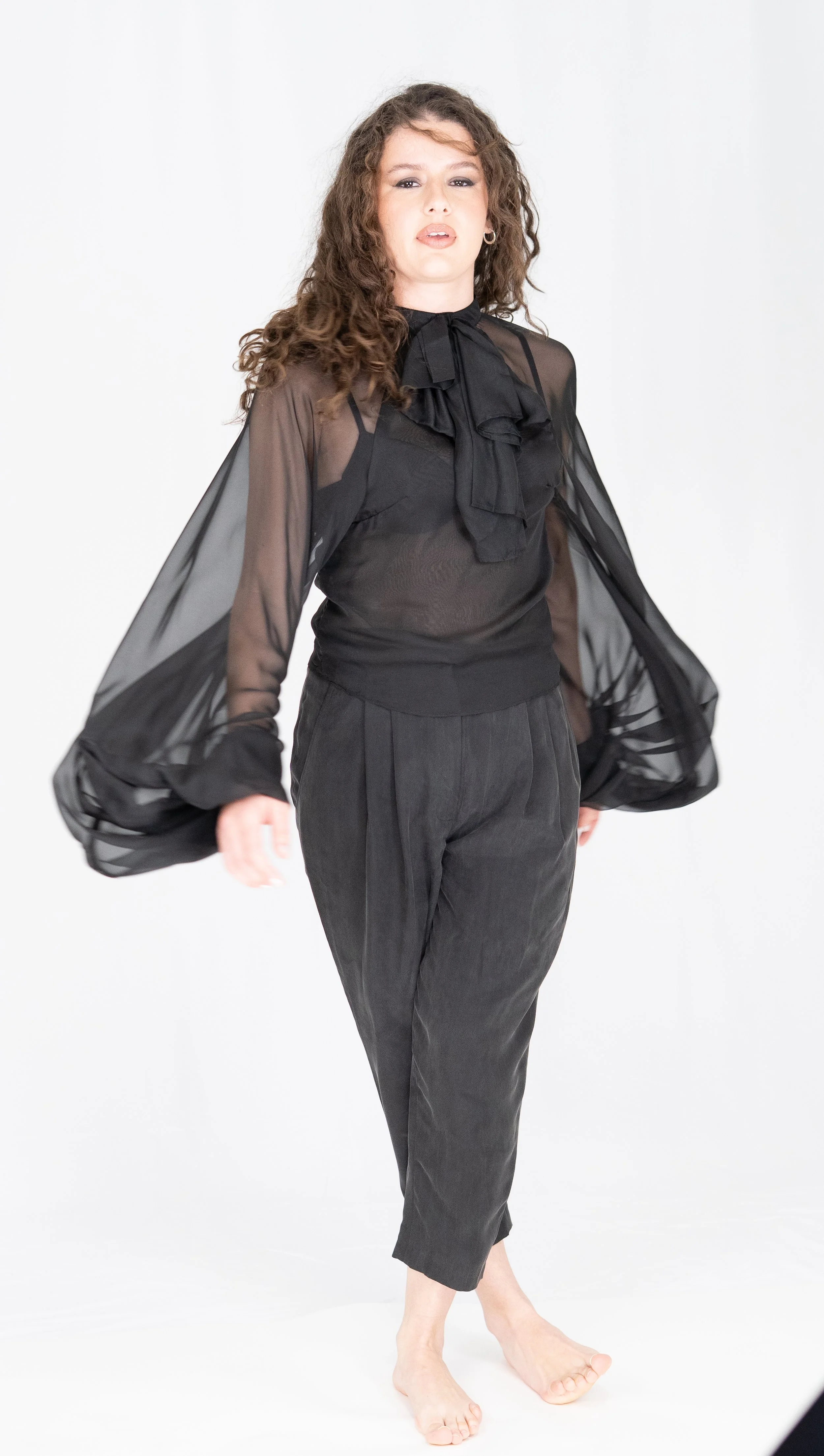 A woman with curly hair in a black sheer blouse with ruffles, black pants, and no shoes standing against a plain white background.