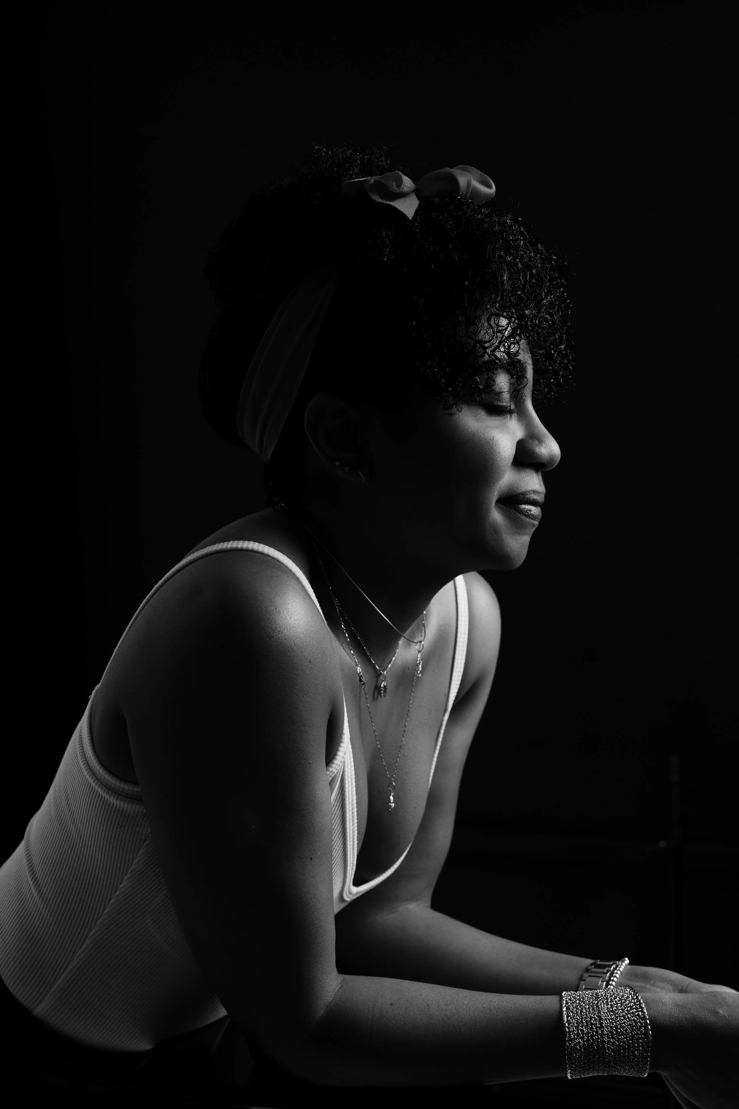 A profile portrait of a woman with curly hair tied with a ribbon, wearing a sleeveless top, layered necklaces, a bracelet, and earrings, captured in black and white with a dark background.