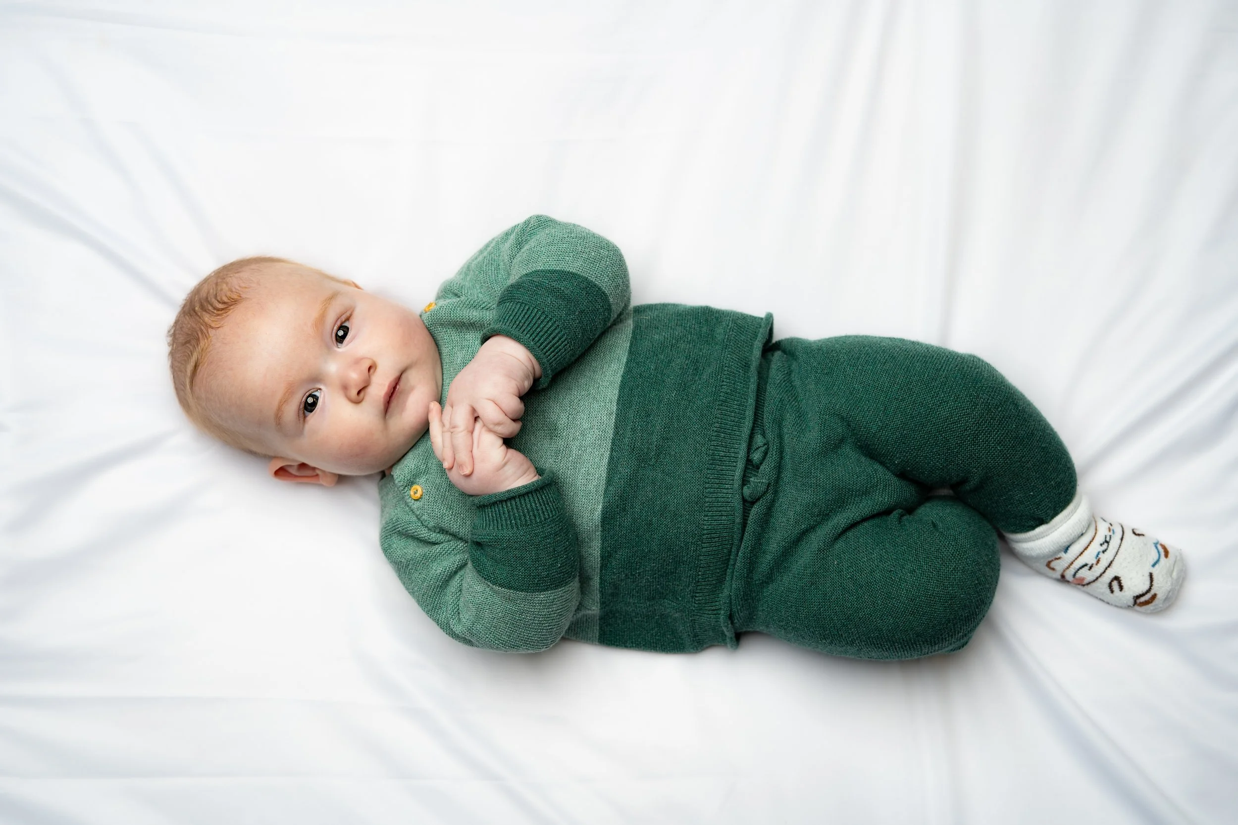 Young child lying on a white sheet, wearing green sweater and pants, with socks featuring a playful pattern.