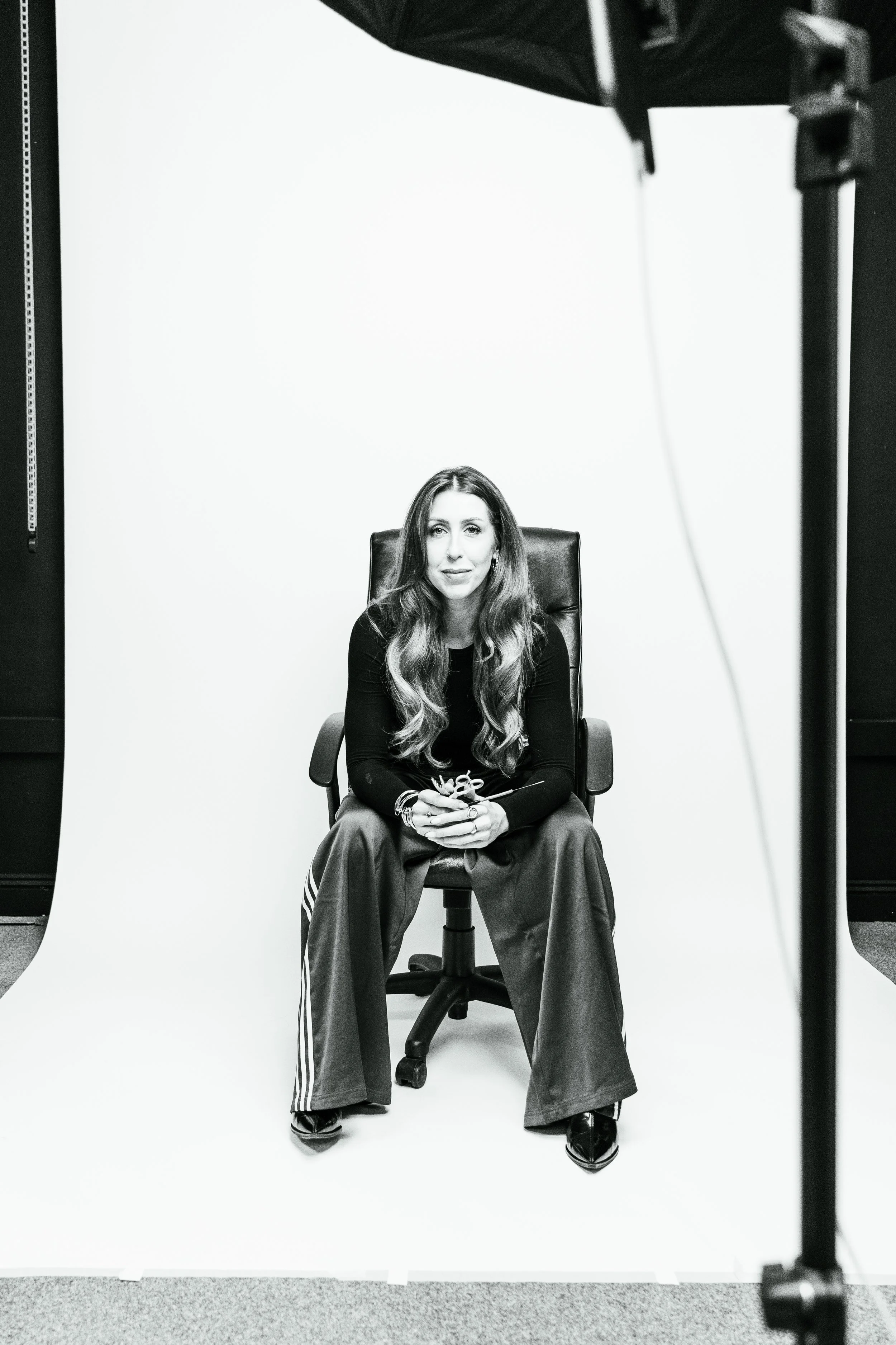 A woman with long hair sitting on a black office chair in front of a white backdrop during a photo shoot.