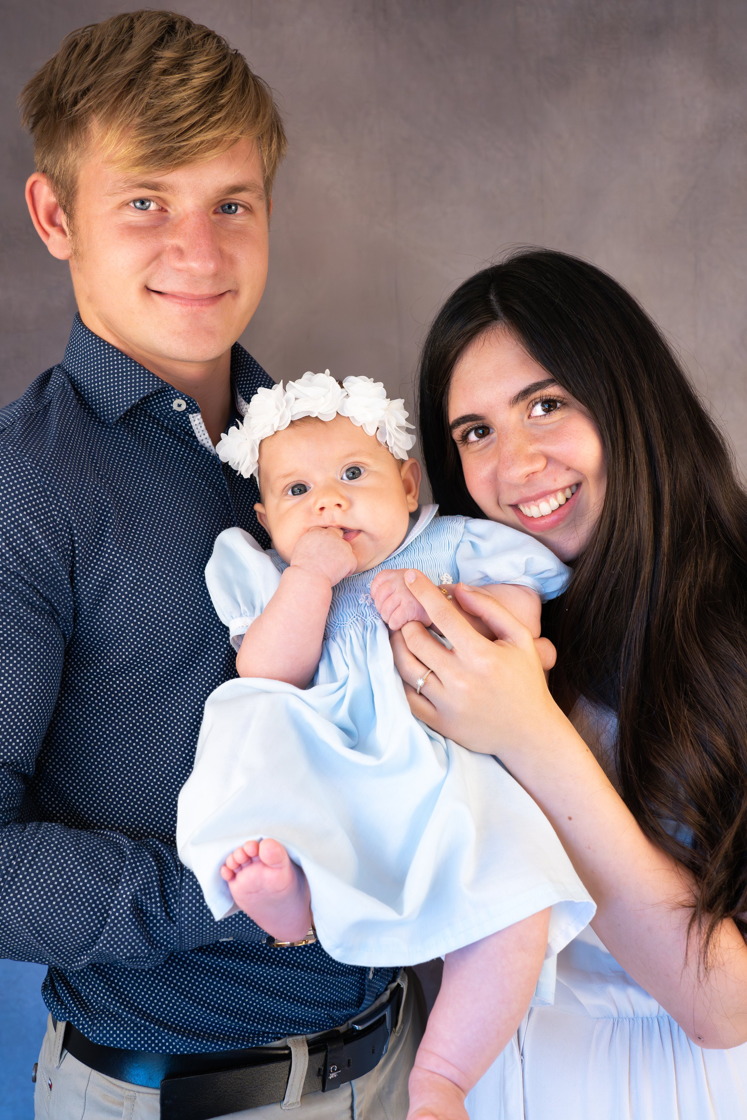 A young couple holding their baby girl, who has a flower headband, against a neutral background.