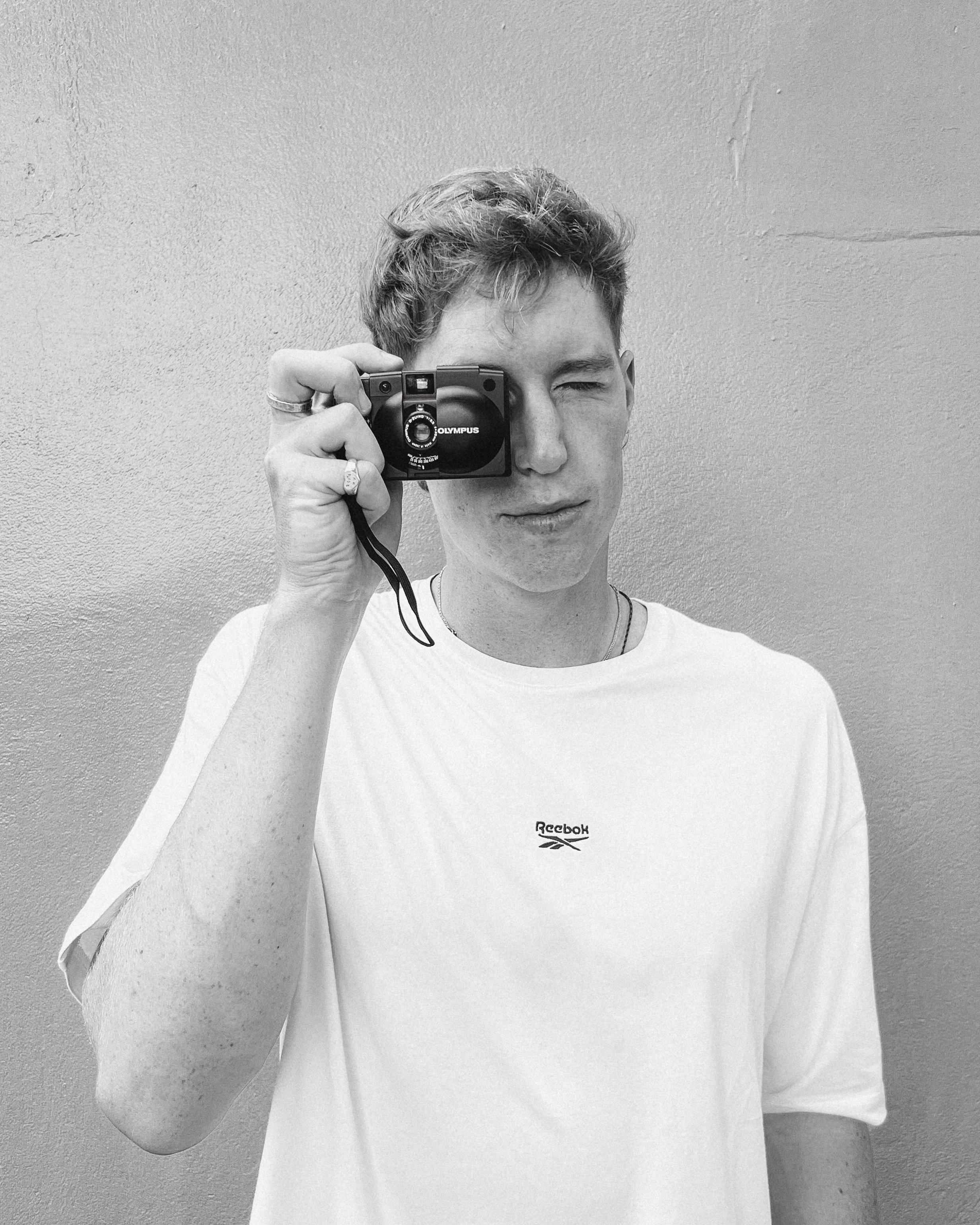 A young man with short wavy hair wearing a Reebok t-shirt, holding a camera up to his face, taking a selfie in front of a plain wall.