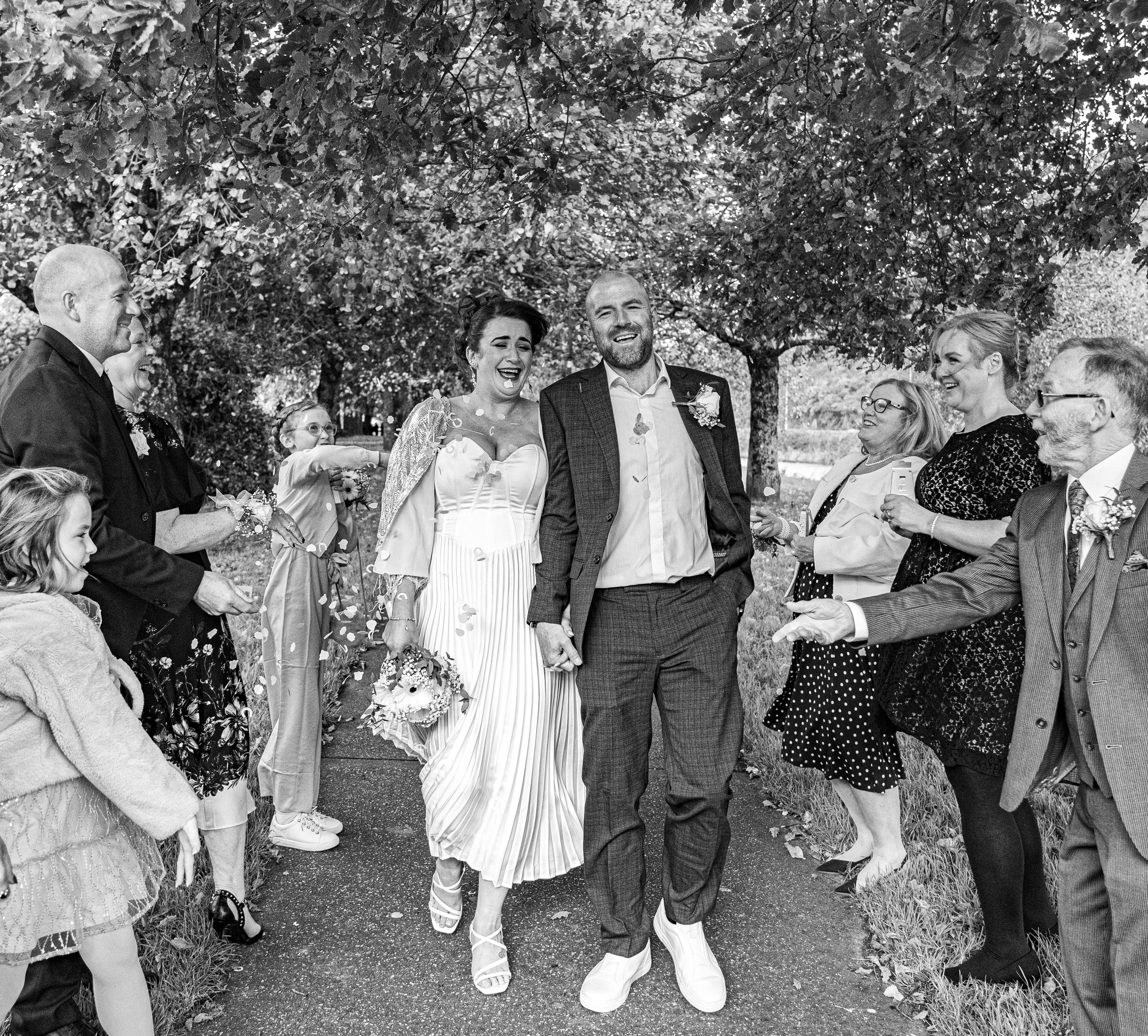 A black and white photo of a newlywed couple walking hand-in-hand through a crowd of family and friends outdoors under trees, celebrating.