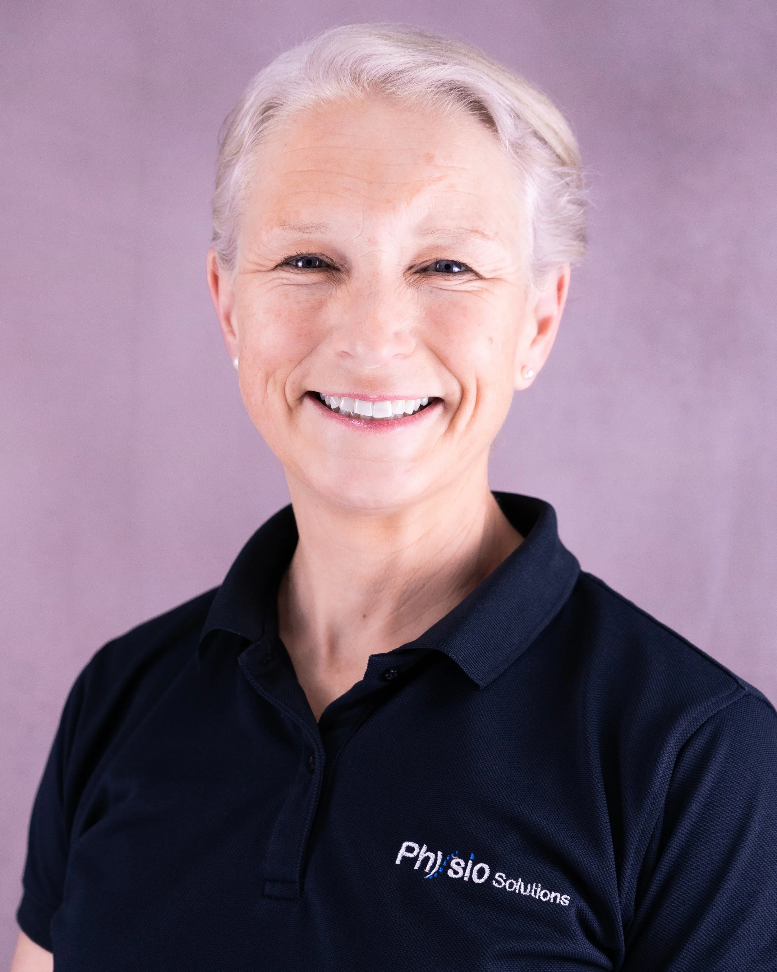 A smiling woman with short, light blonde hair, wearing a navy blue polo shirt with the logo 'Phys.io Solutions' on it, standing against a soft purple background.