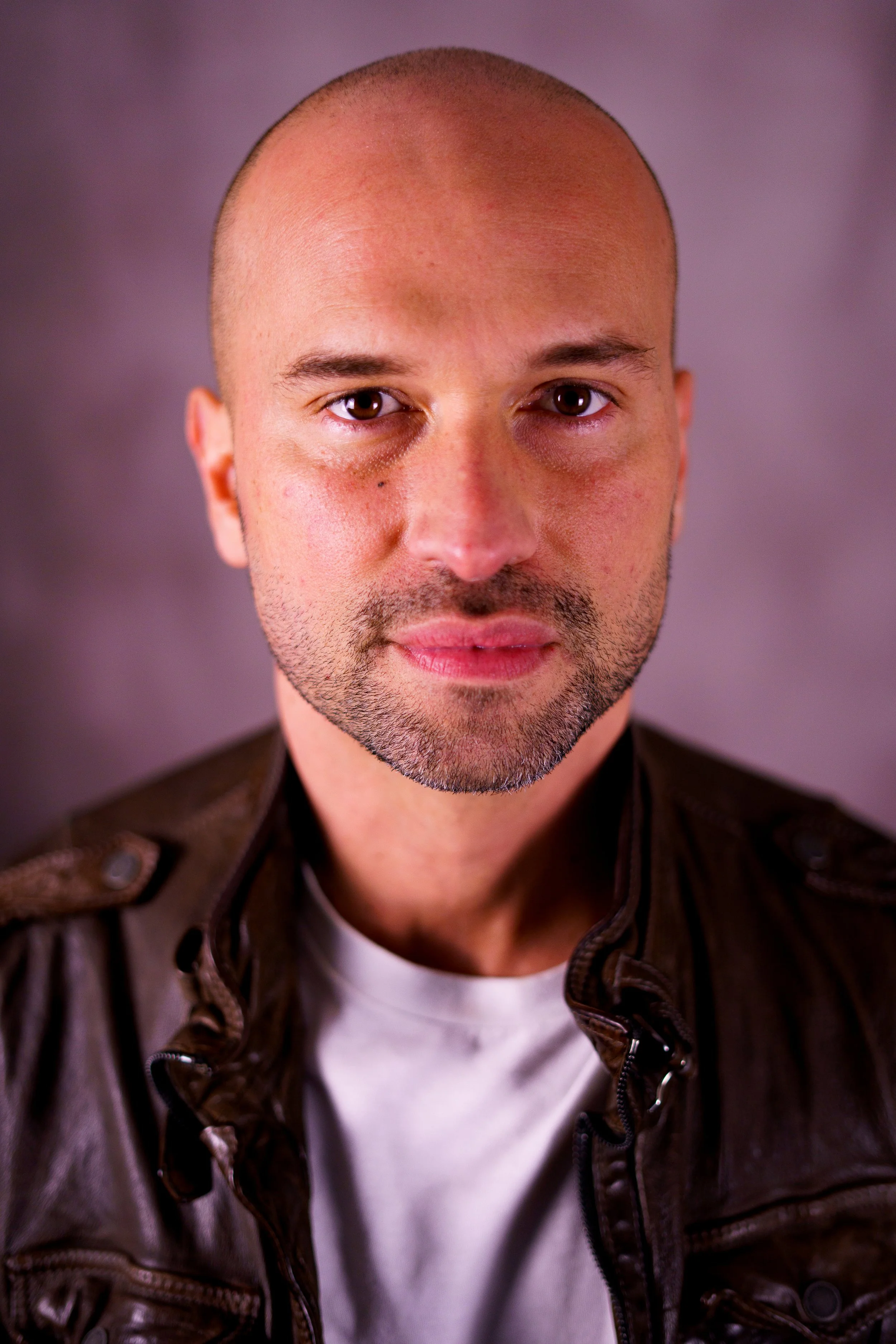 Close-up portrait of a bald man with a short beard, wearing a leather jacket over a white t-shirt, looking directly at the camera with a neutral expression.