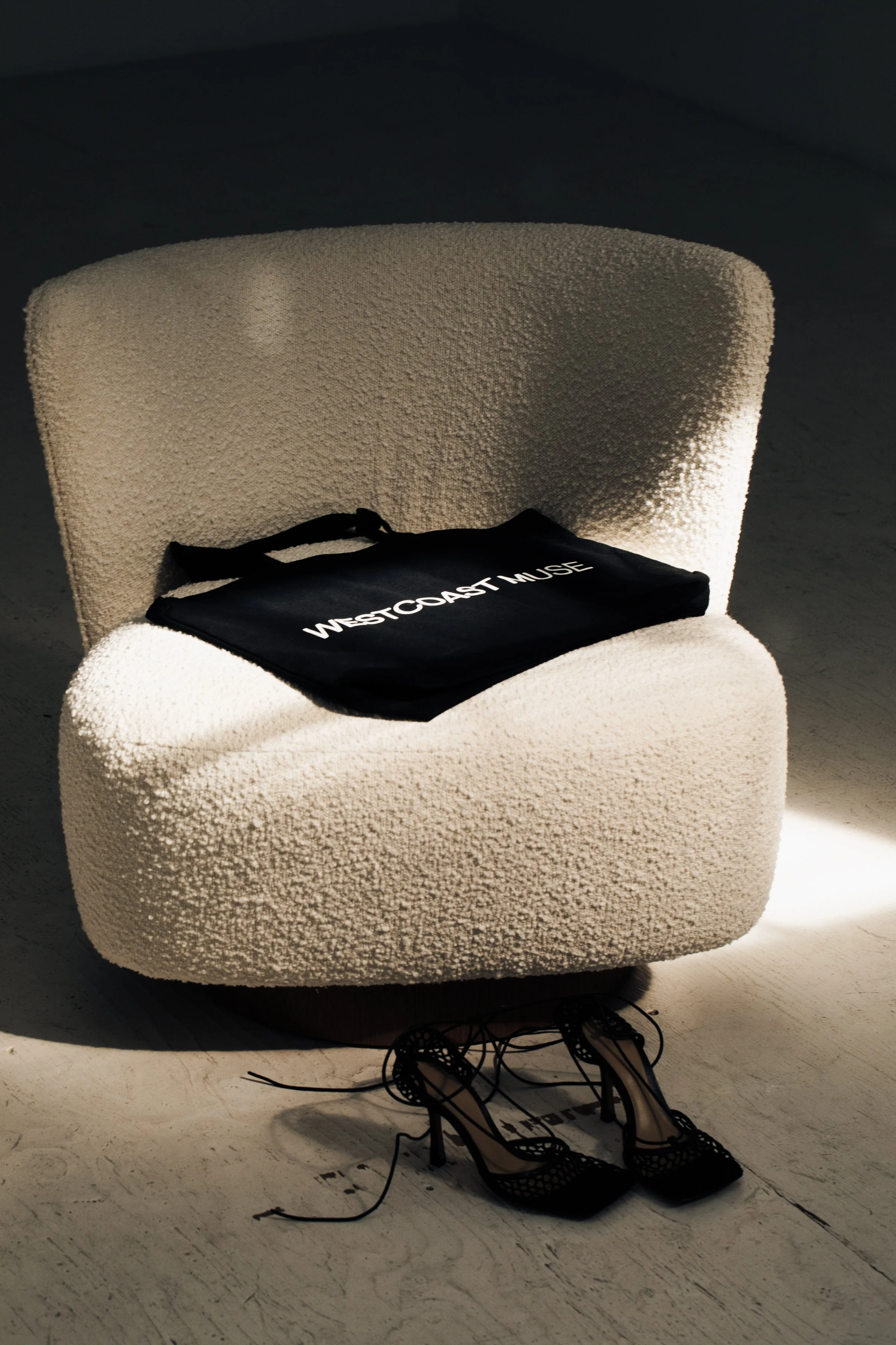 A white textured armchair with a black tote bag labeled 'West Coast Muse' on it, and a pair of black high-heeled shoes with cutout details placed on the floor in front of the chair.