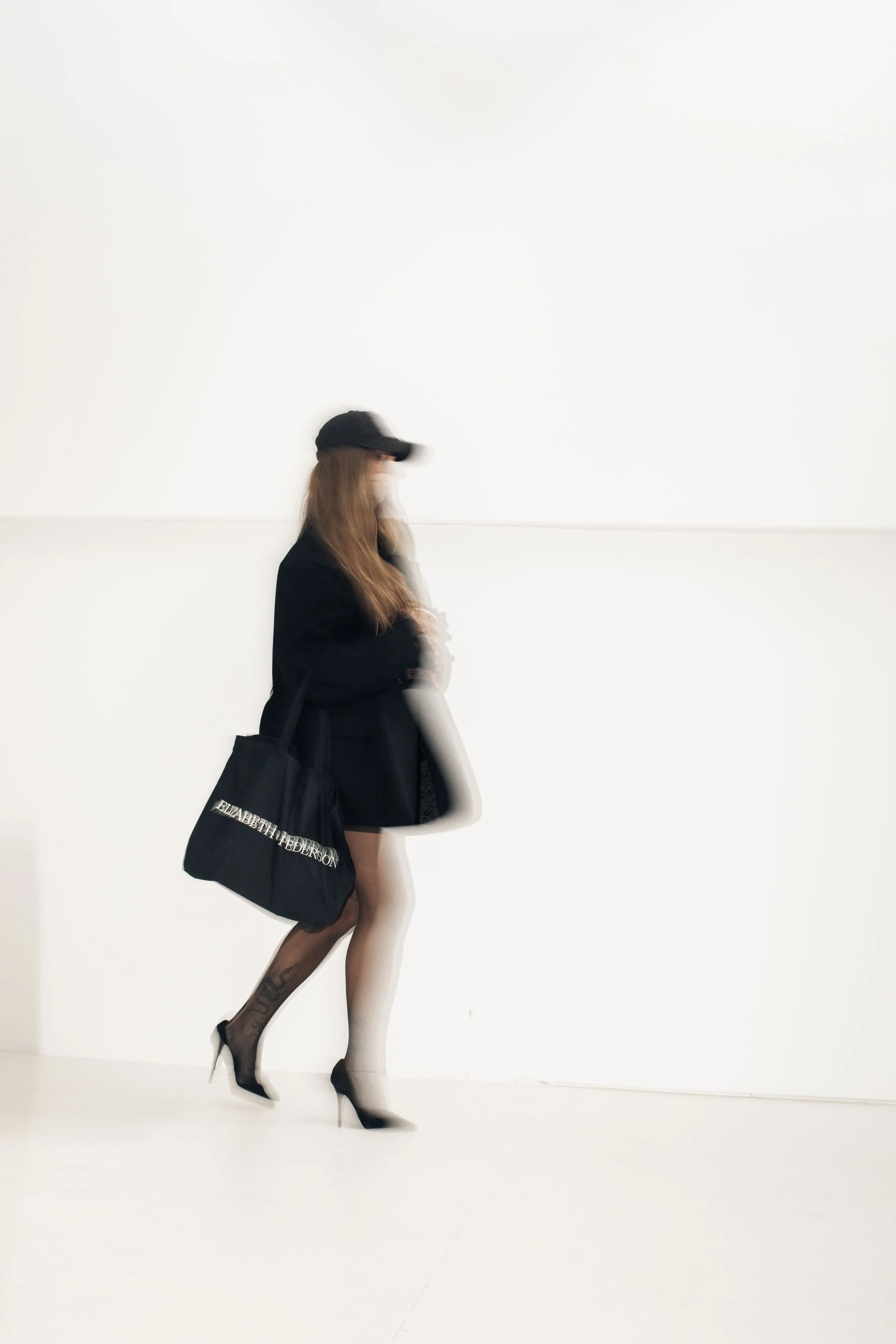 A woman walking in a plain white space wearing high heels, a black blazer, black cap, with a black tote bag and transparent hosiery.