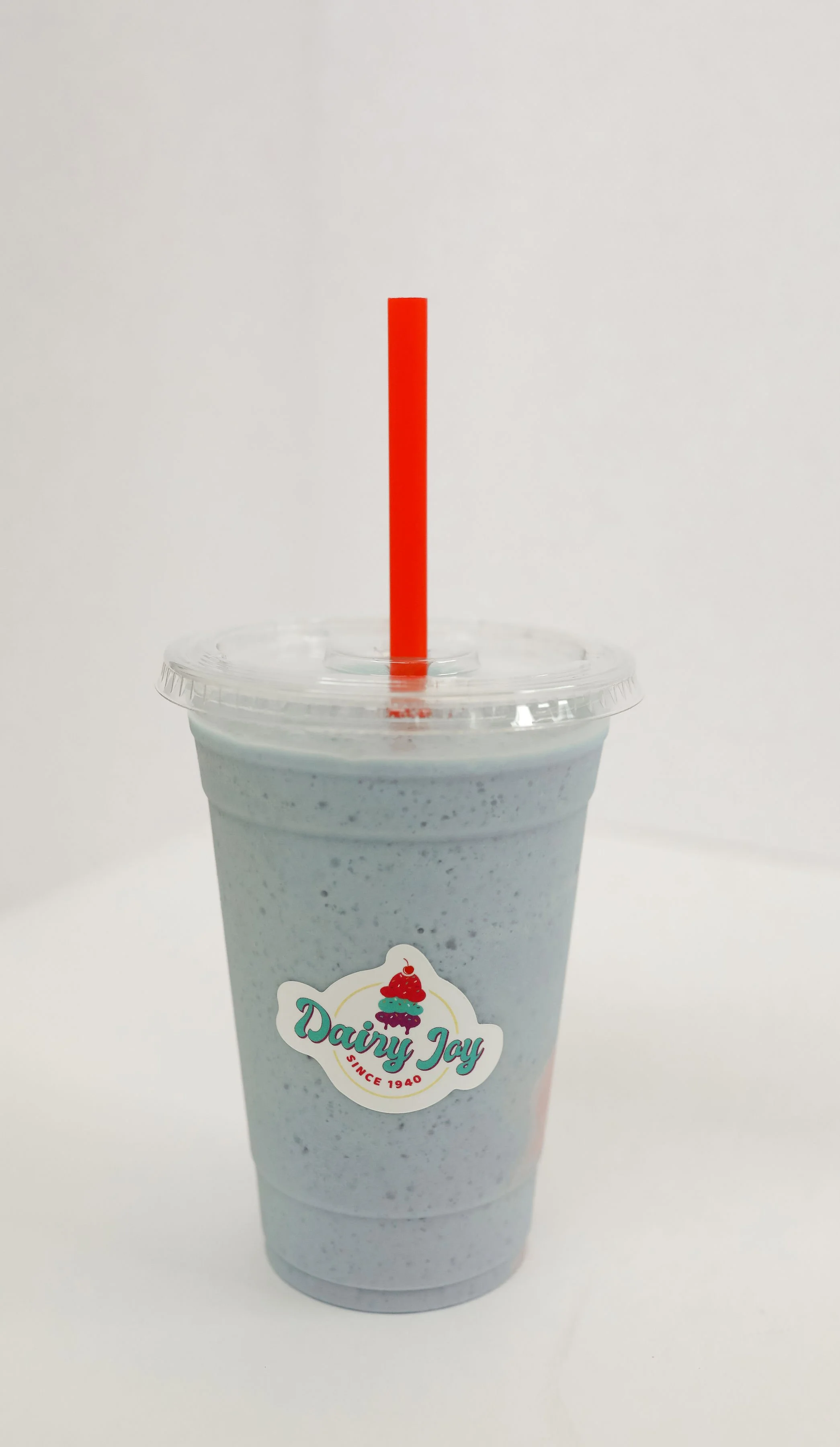 A milkshake in a clear plastic cup with a domed lid and a red straw. The milkshake is light gray with small dark specks. The cup has a Dairy Joy logo with a colorful ice cream cone graphic and the text "Dairy Joy Since 1940." The background is plain white.
