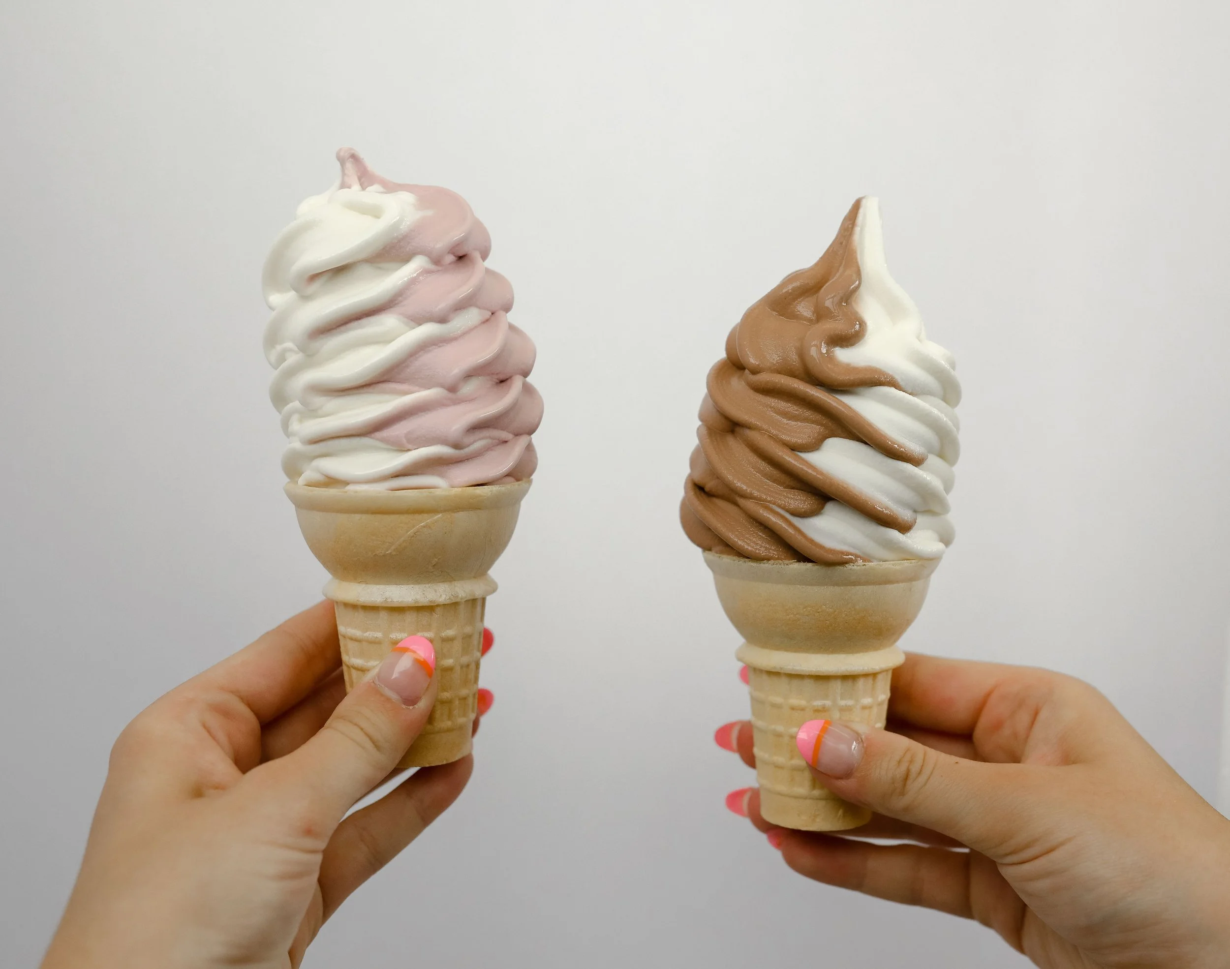 Two hands holding ice cream cones with soft serve ice cream, one with pink and white swirls and the other with brown and white swirls, against a plain white background.