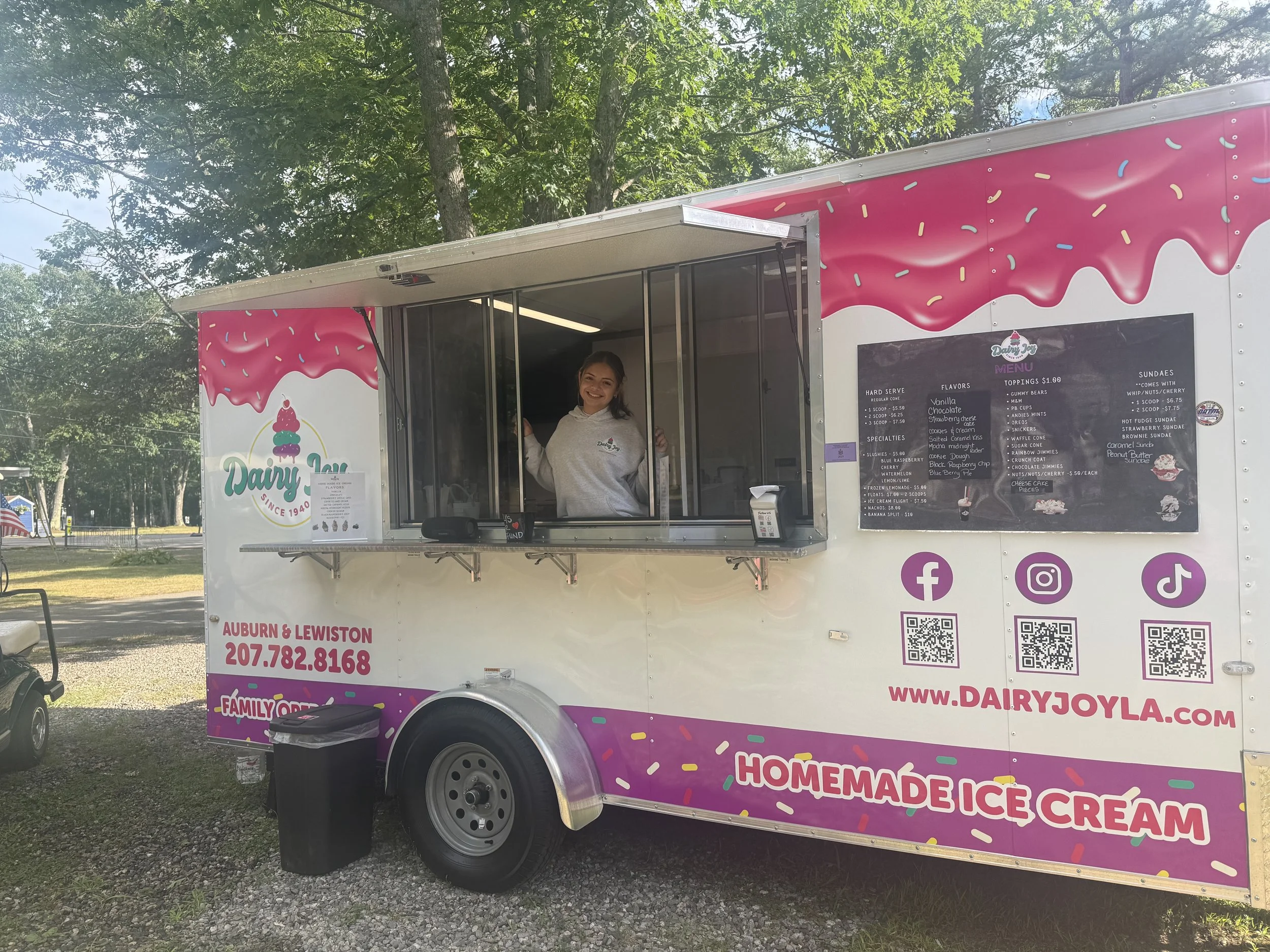Pink ice cream truck with colorful sprinkles design, woman smiling and standing at the window, menu listing flavors and toppings, social media icons, QR codes, and contact information on the truck, trees in the background.
