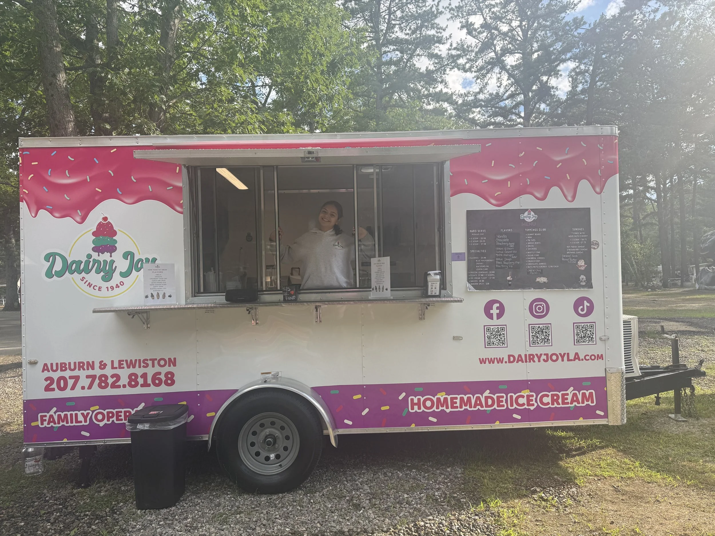 A pink and white ice cream truck themed with sprinkles and ice cream graphic, parked outdoors with trees and a blue sky. A smiling woman stands inside the window, serving ice cream.